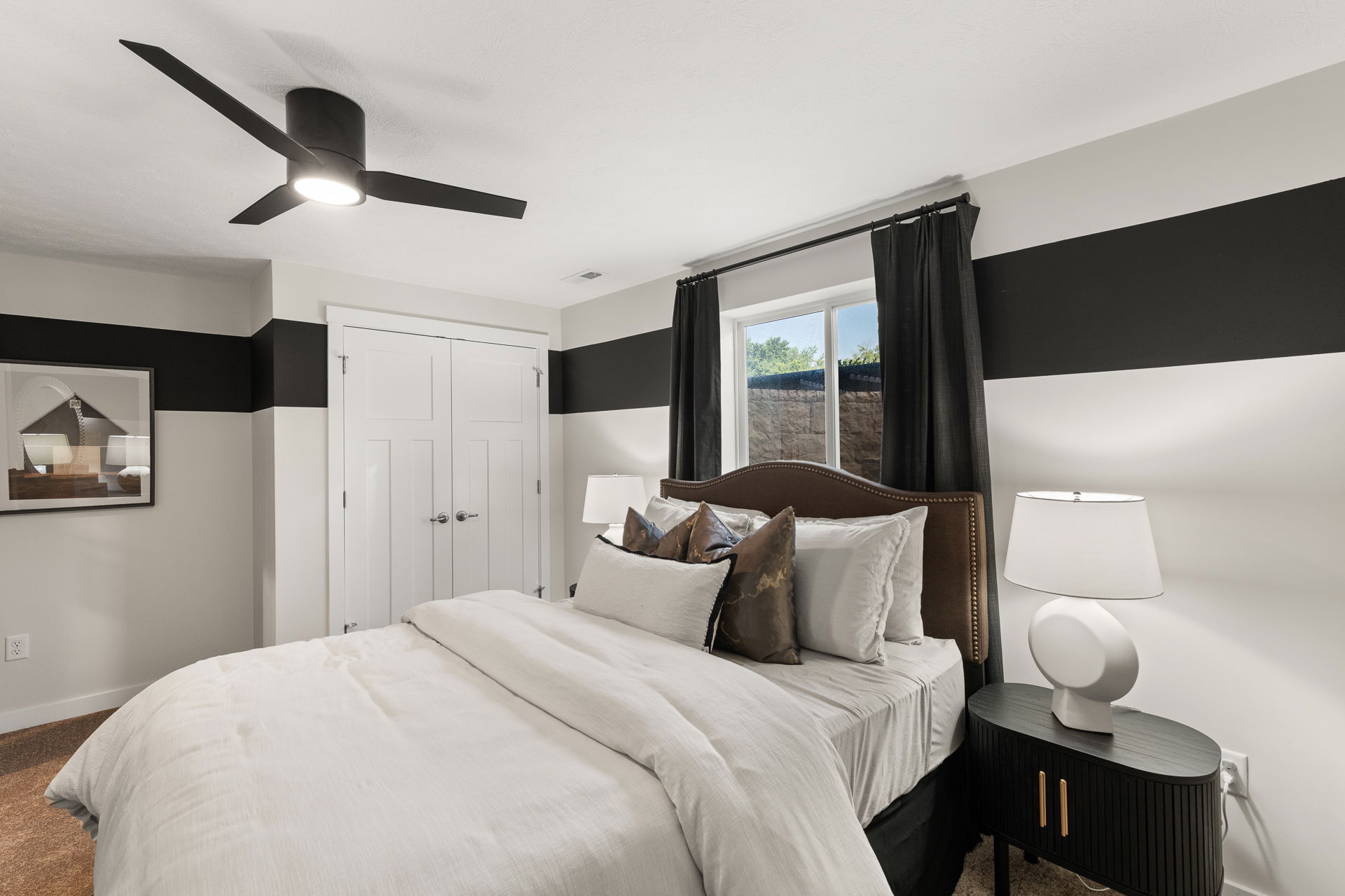 Modern bedroom with a cozy bed, stylish black and white decor, ceiling fan, and natural light from a window.