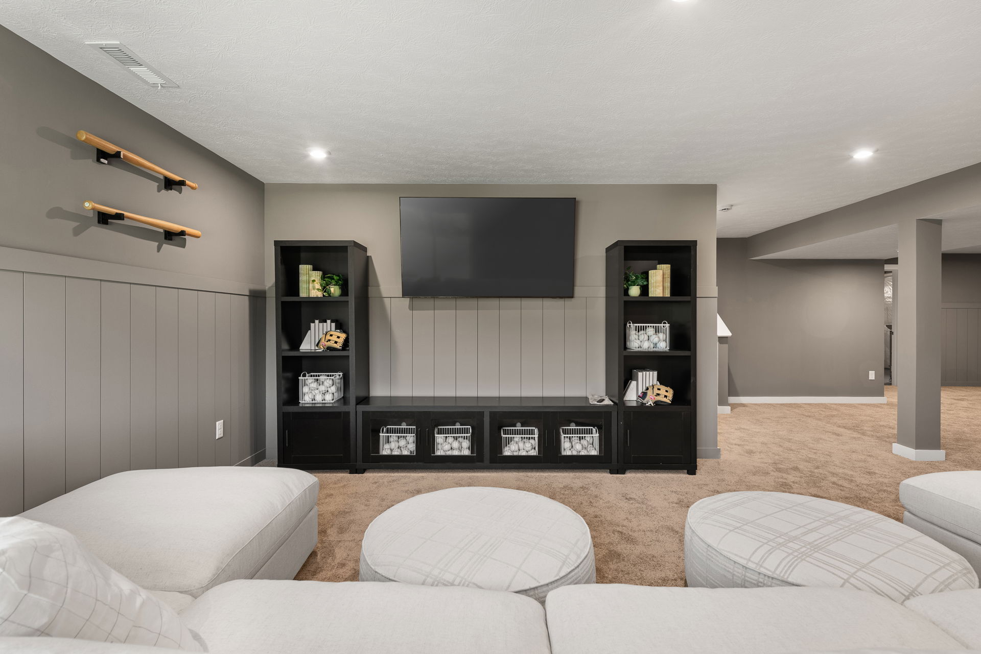 A modern basement entertainment area featuring a large wall-mounted TV, black shelving units, and a cozy beige sectional sofa.