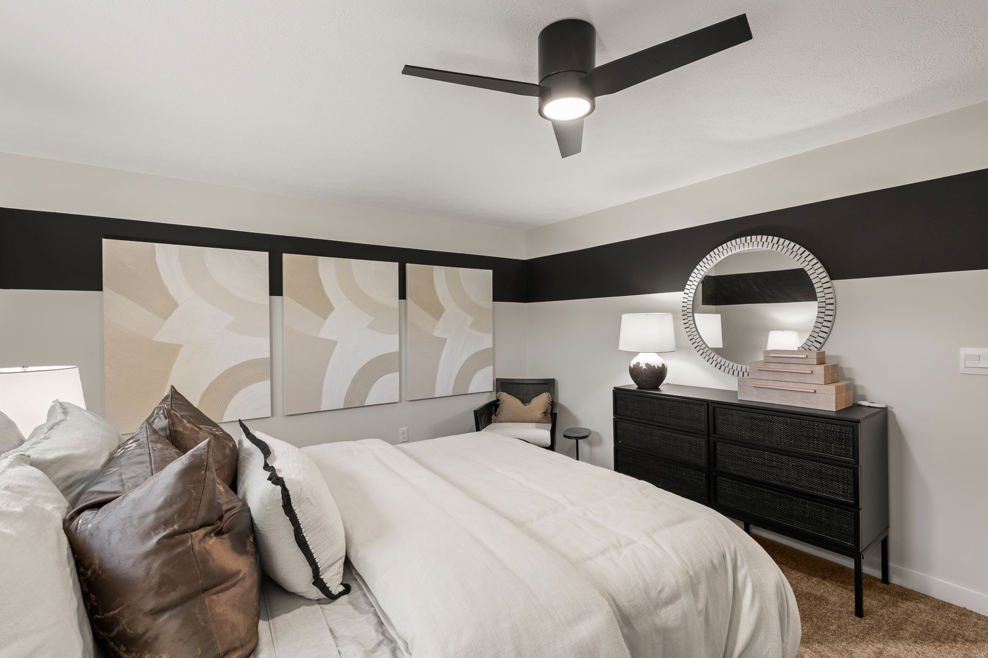 Modern bedroom with stylish decor, featuring a black ceiling fan, geometric wall art, and a sleek dresser with a round mirror.