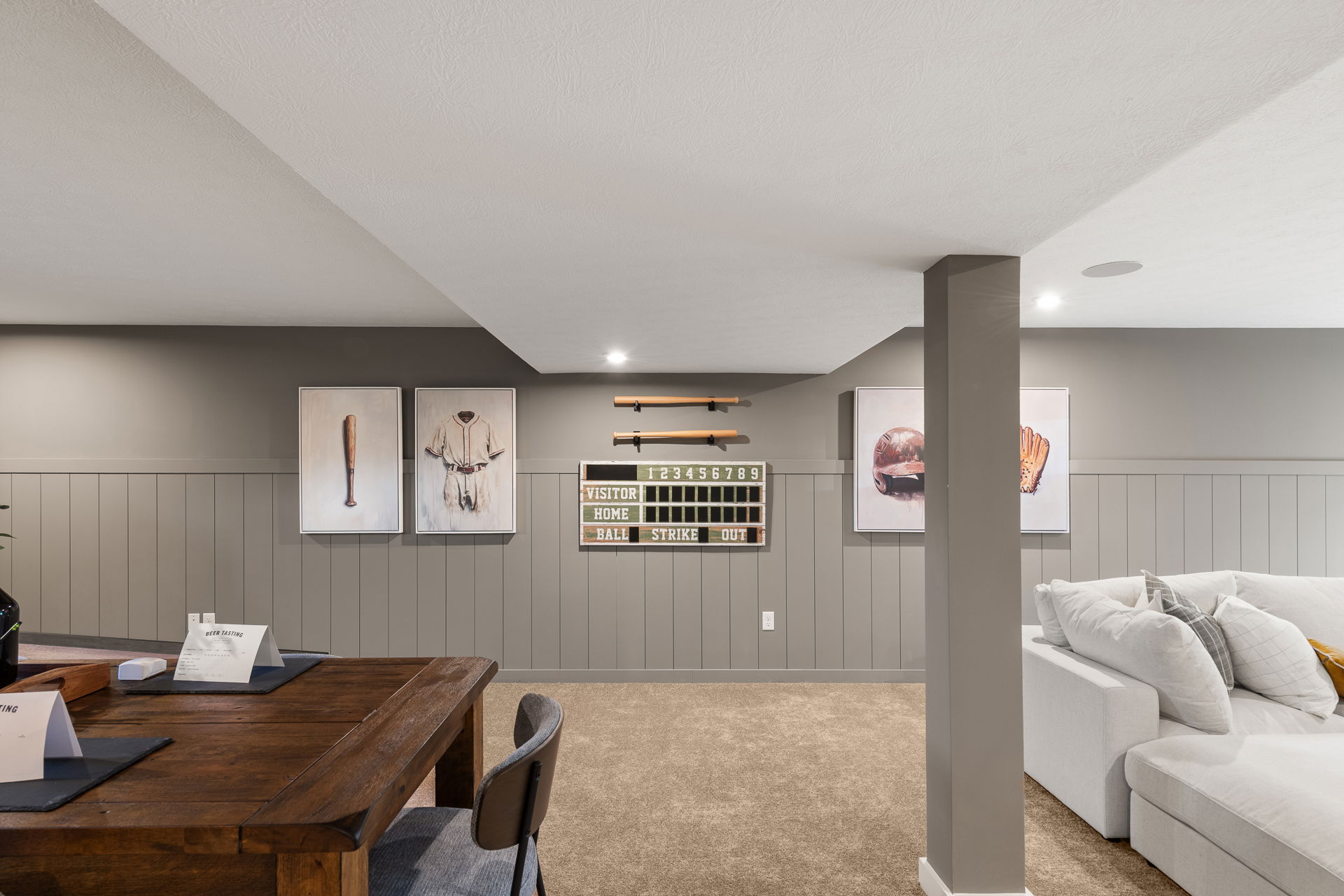 Modern basement recreation room with baseball-themed wall art, gray wainscoting, and cozy seating area.