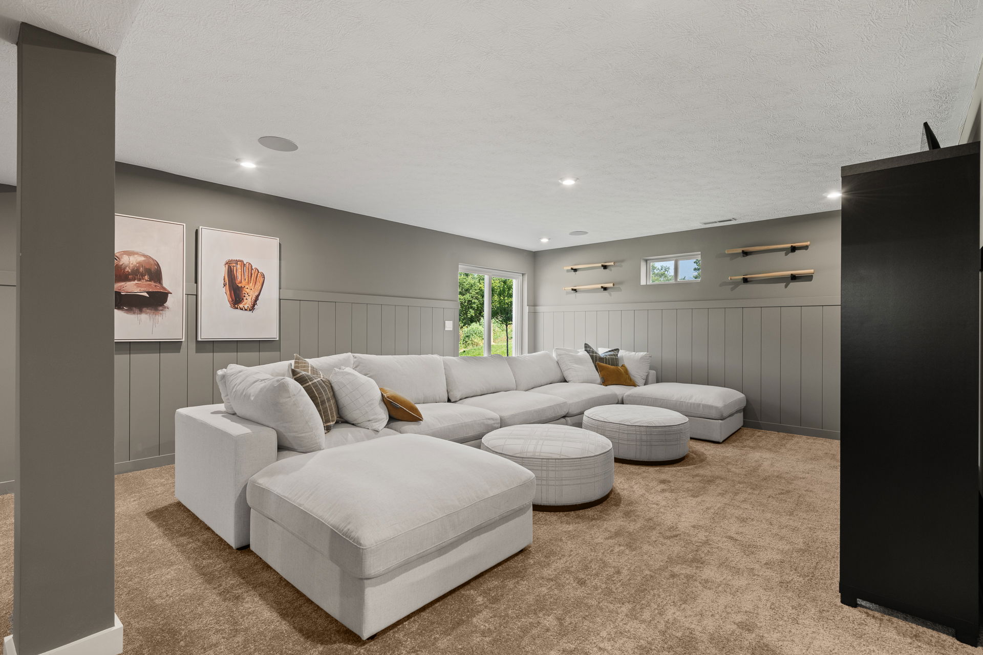 Stylish basement family room with large sectional sofa, cozy carpet, sports-themed artwork, and ample natural light.