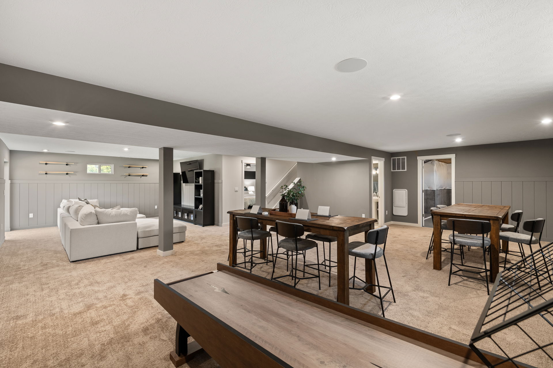 Modern finished basement with beige carpet, comfortable sectional sofa, wall-mounted TV, shuffleboard table, and wooden dining tables.