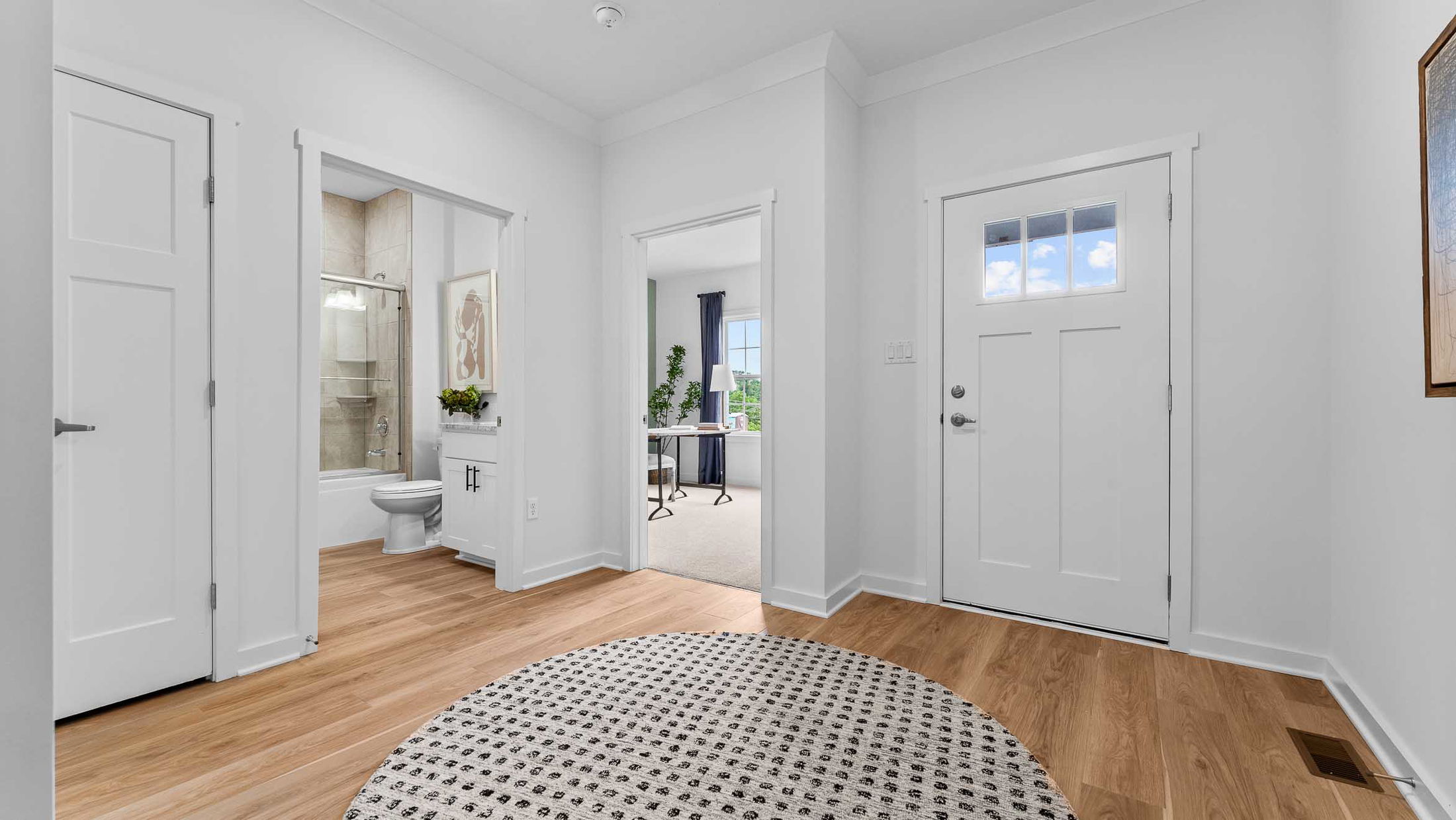 Bright, modern home entryway featuring a wooden floor, textured rug, bathroom, and office space.