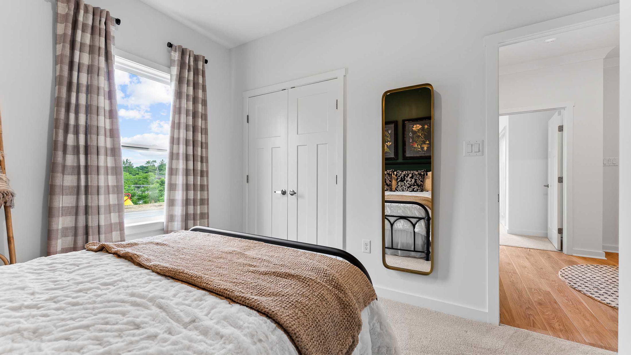 Bright and cozy bedroom with a plaid curtain, fluffy white bedspread, wall mirror, and wood accents creating a serene atmosphere.