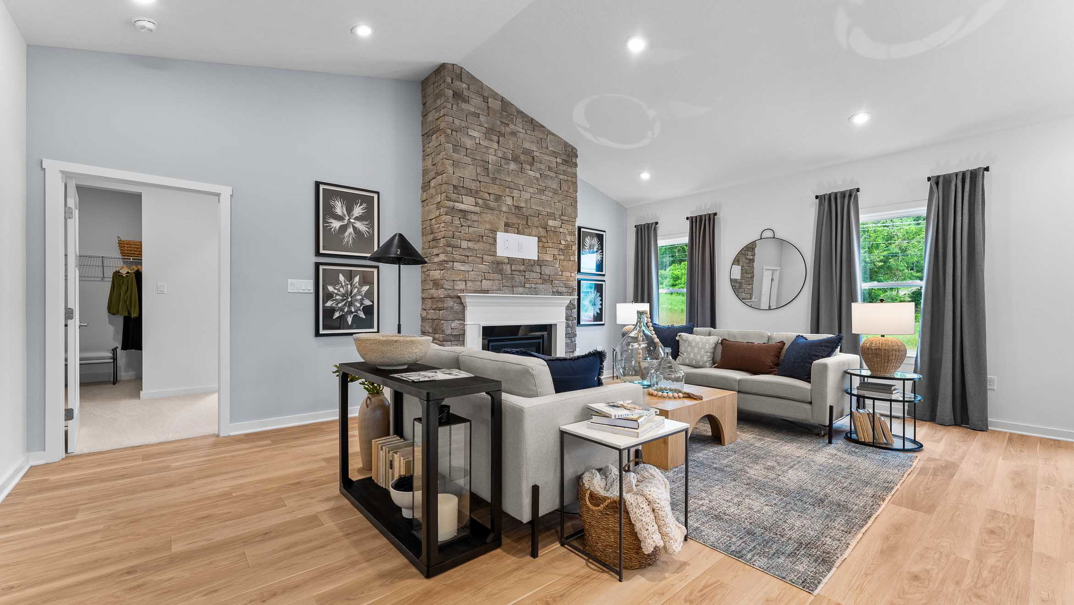 Modern living room with stone fireplace, stylish gray sofas, wooden flooring, and large windows showcasing a lush green view.