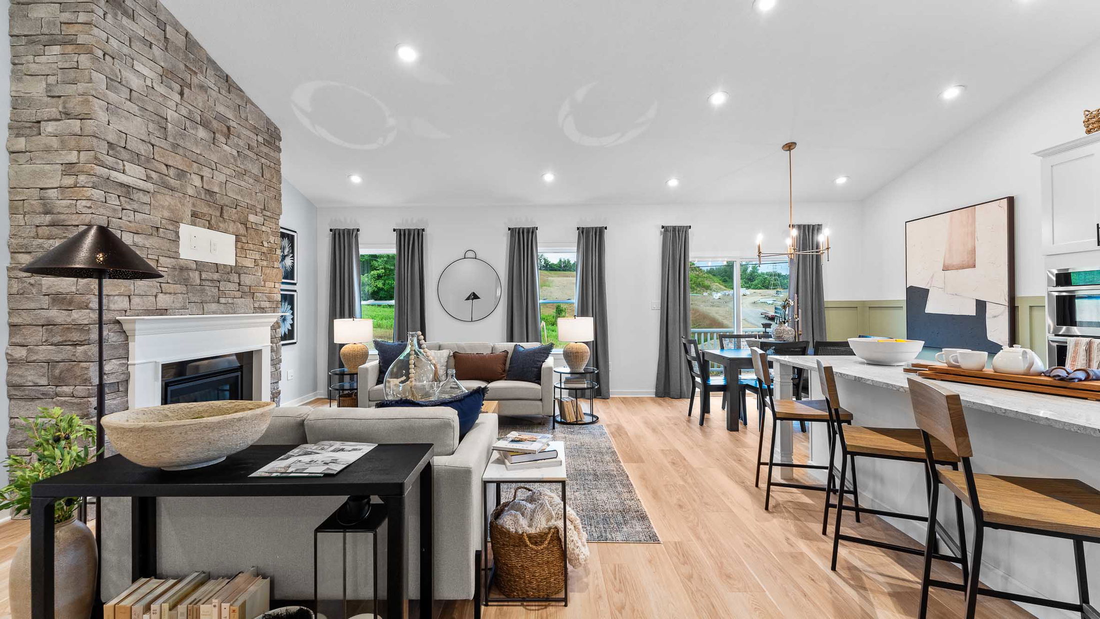 Modern open-concept living room and kitchen with stone fireplace, neutral decor, and wooden flooring.