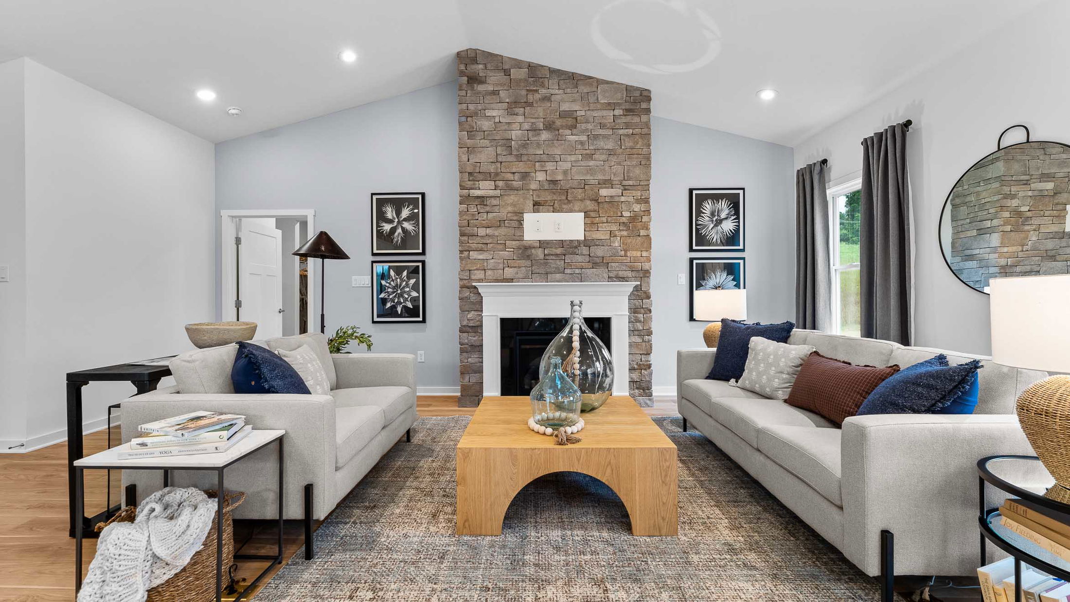 A modern living room with a stone fireplace, gray sofas, and a wooden coffee table featuring decorative glass jars, accented by contemporary art and neutral tones.