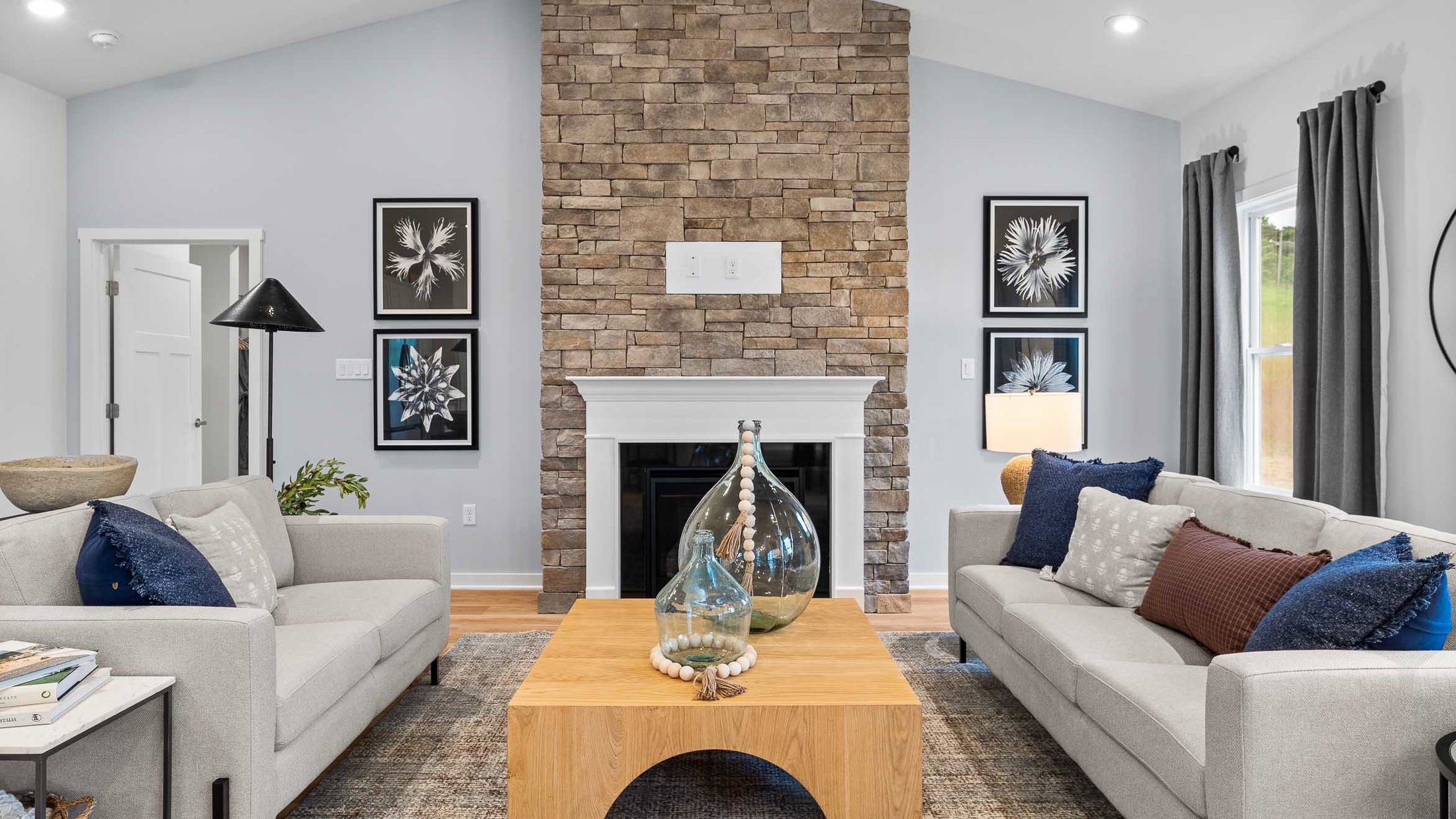 Modern living room with a stone fireplace, neutral-tone sofas, decorative vases on a wooden coffee table, and monochrome floral artwork.
