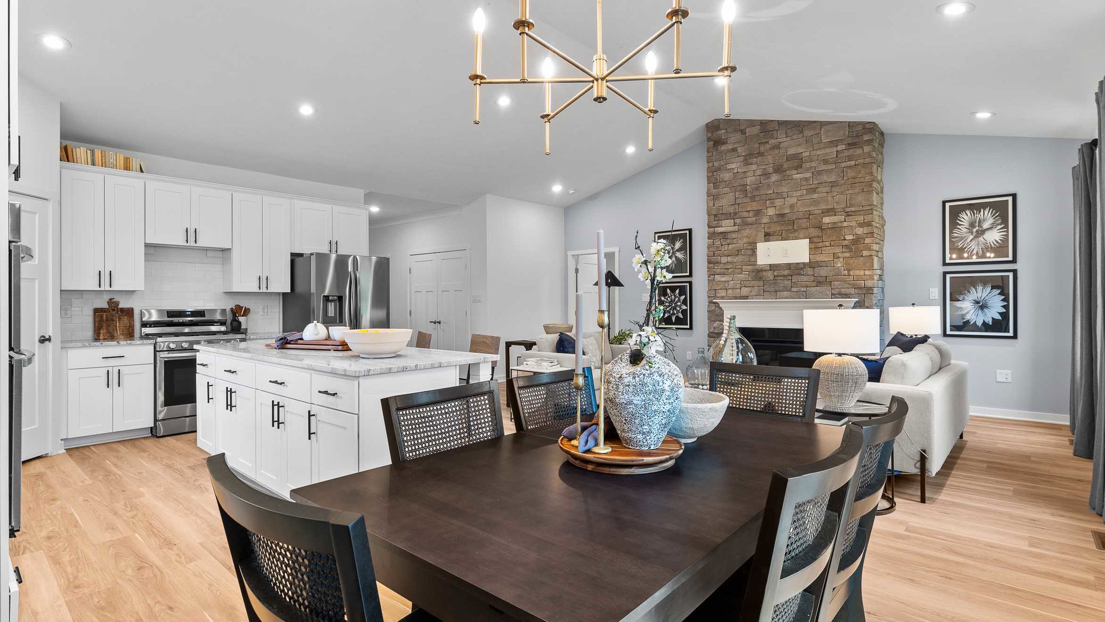 Modern open-concept kitchen and living room with white cabinetry, stainless steel appliances, stone fireplace, and elegant dining area.