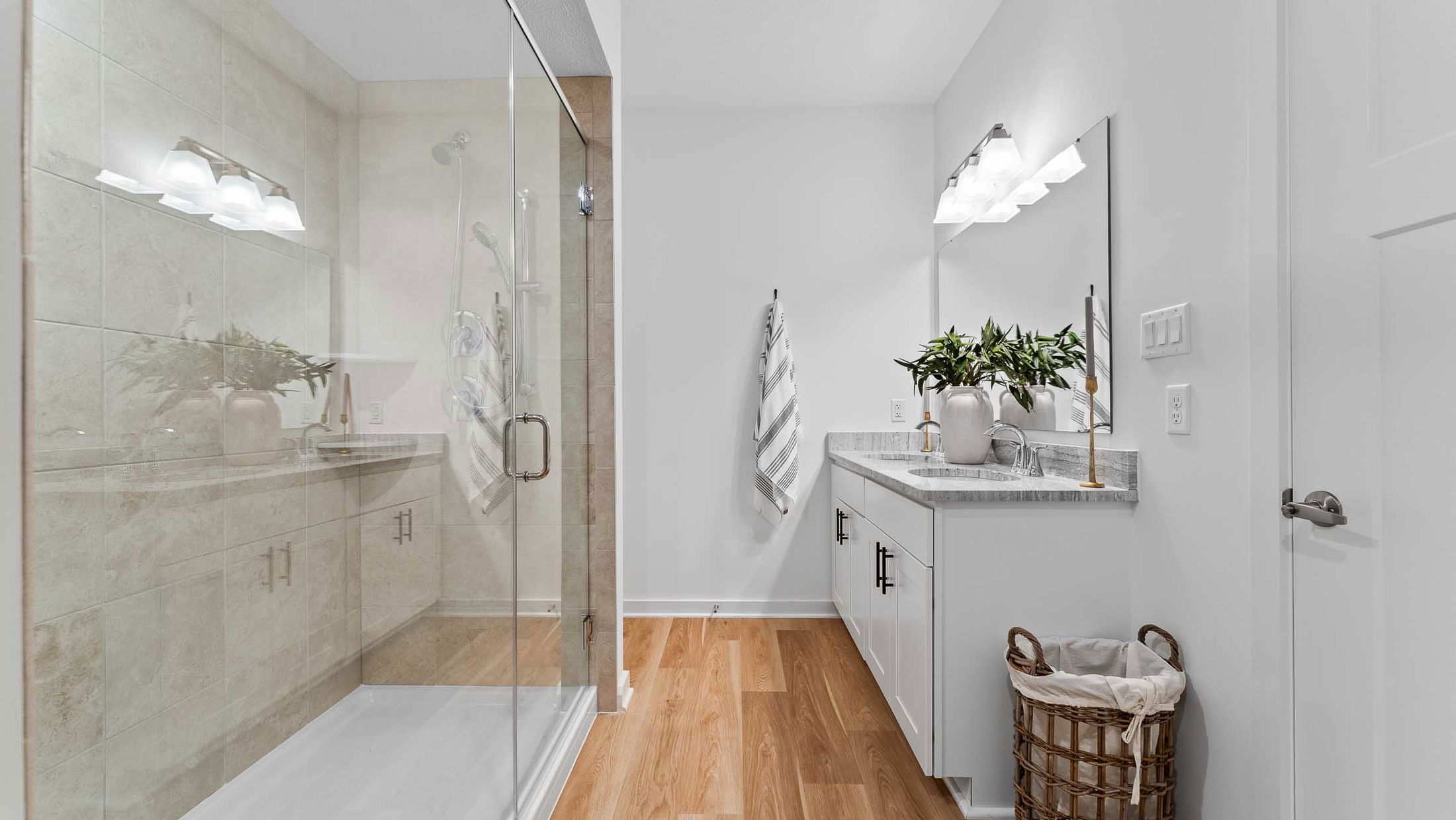 Modern bathroom with glass shower enclosure, white cabinetry, and wooden flooring, featuring plants and neatly folded towels.
