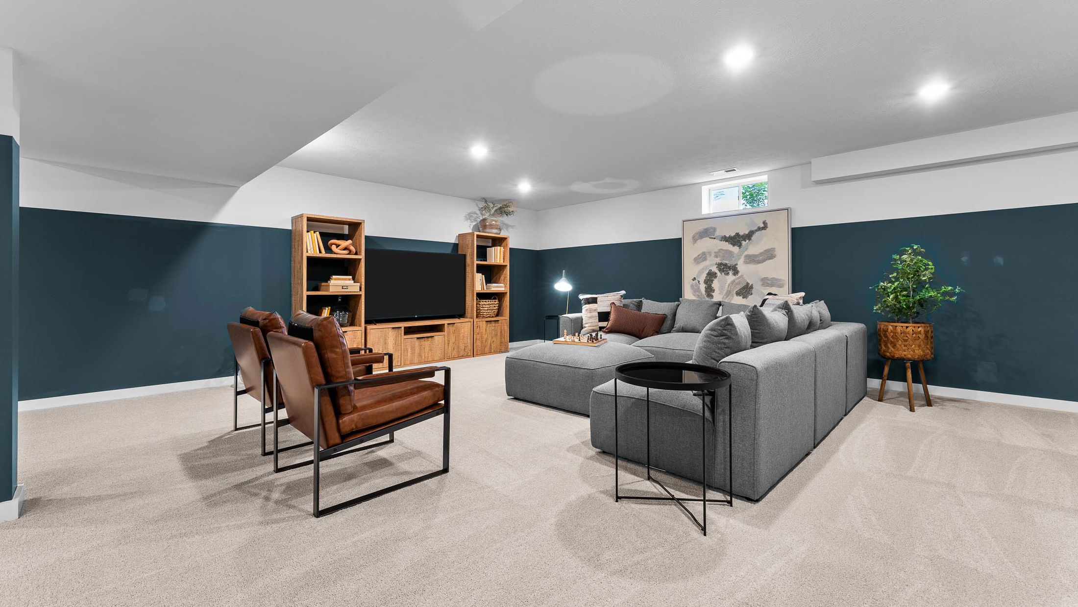 Stylish basement living room with modern gray sectional sofa, flat-screen TV, leather chairs, and wooden shelving.