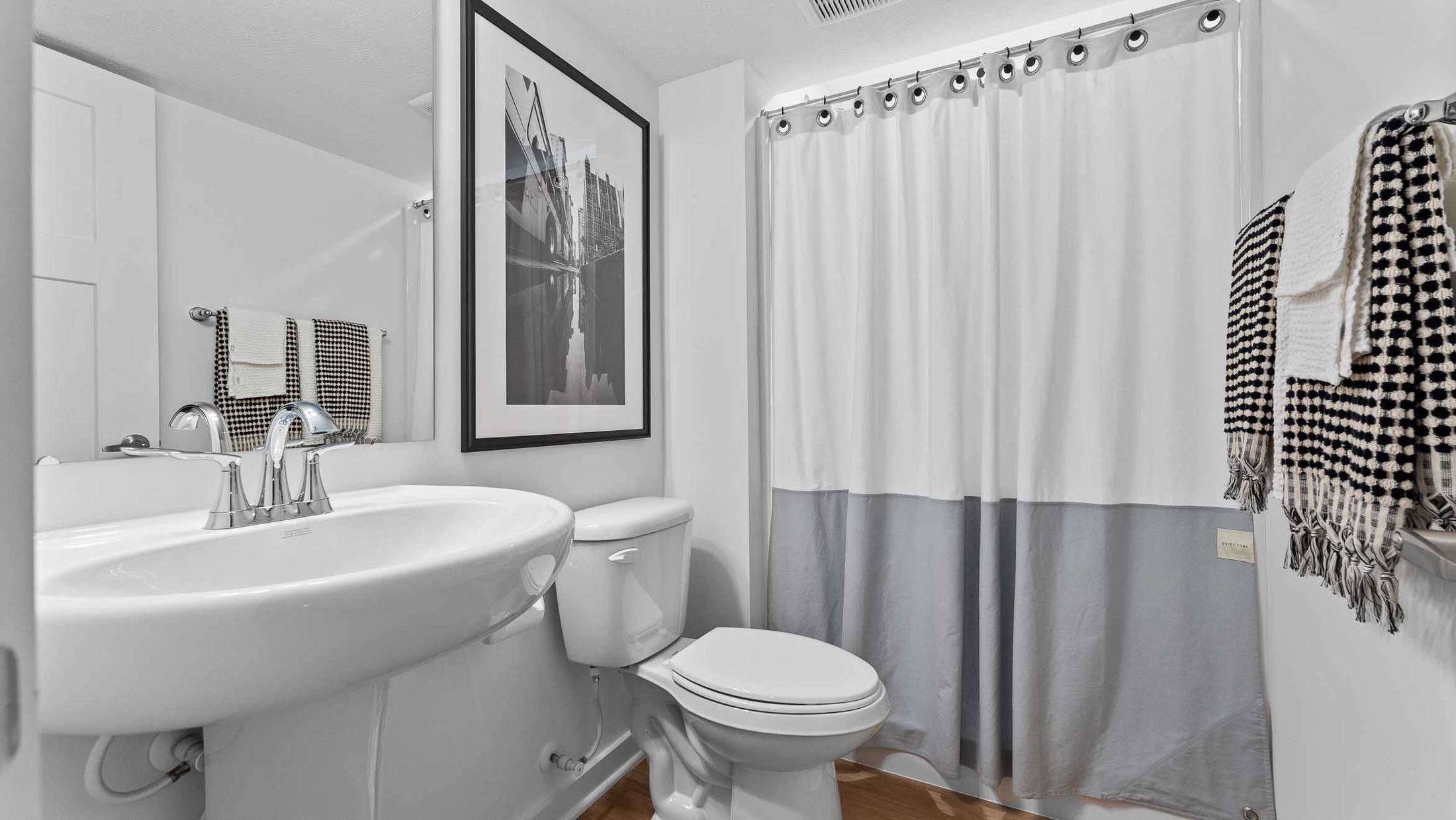 Modern white bathroom with pedestal sink, toilet, large framed wall art, and a shower curtain decorated with black and white towels.