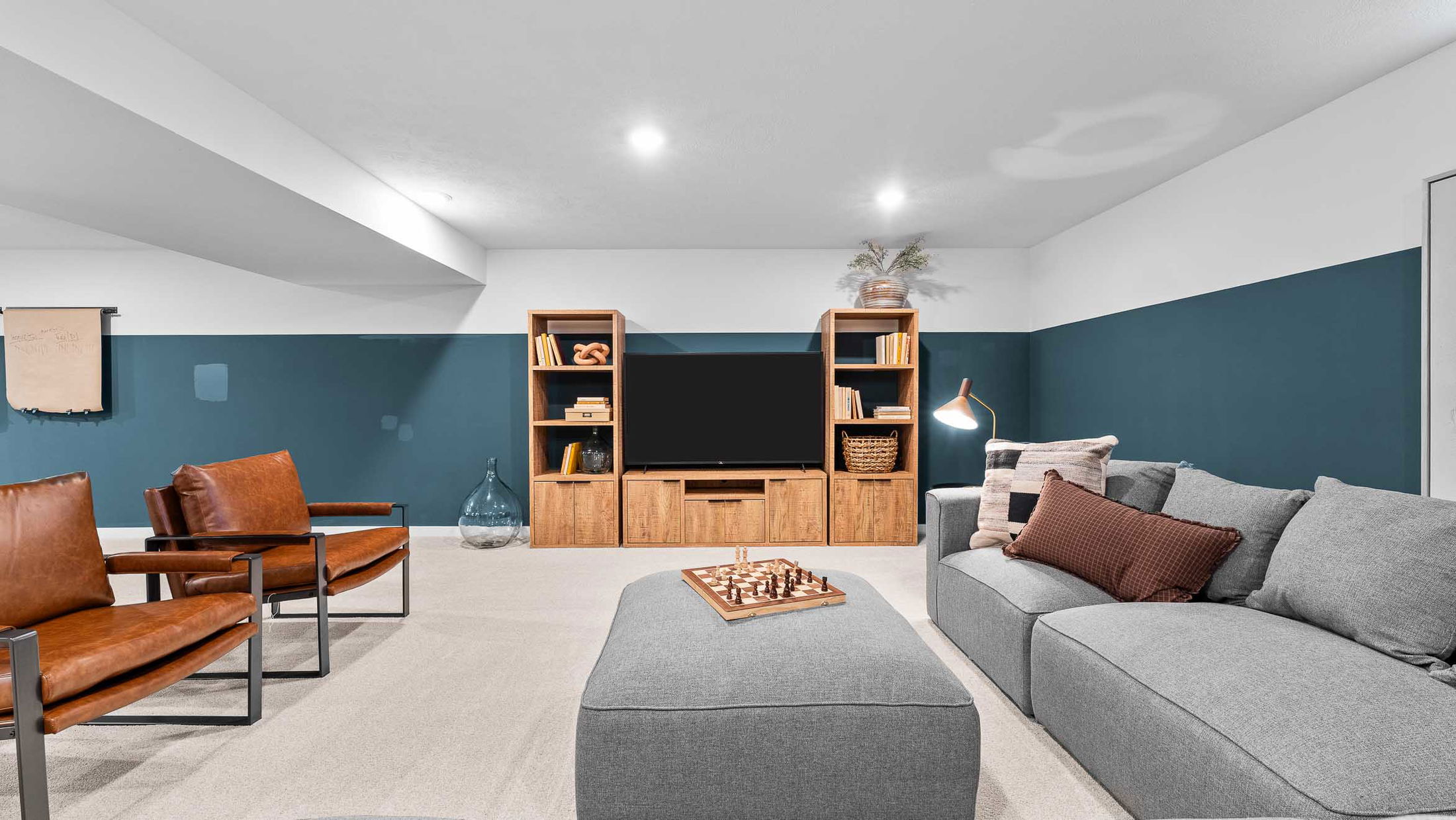 Modern living room with gray sectional sofa, wooden media center, leather chairs, and a chess board on a cushioned ottoman.
