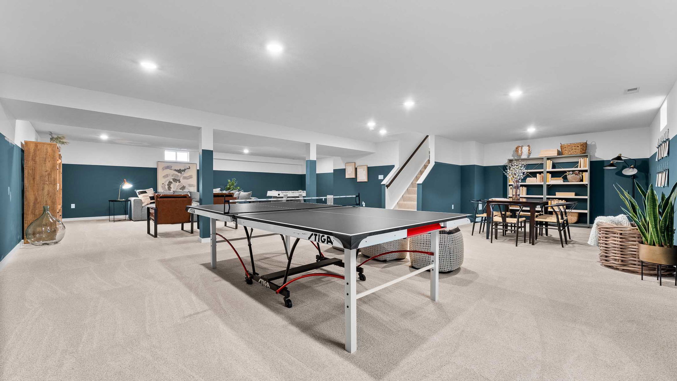 Spacious finished basement with modern decor, featuring a ping pong table, seating area, and bookshelf under bright recessed lighting.