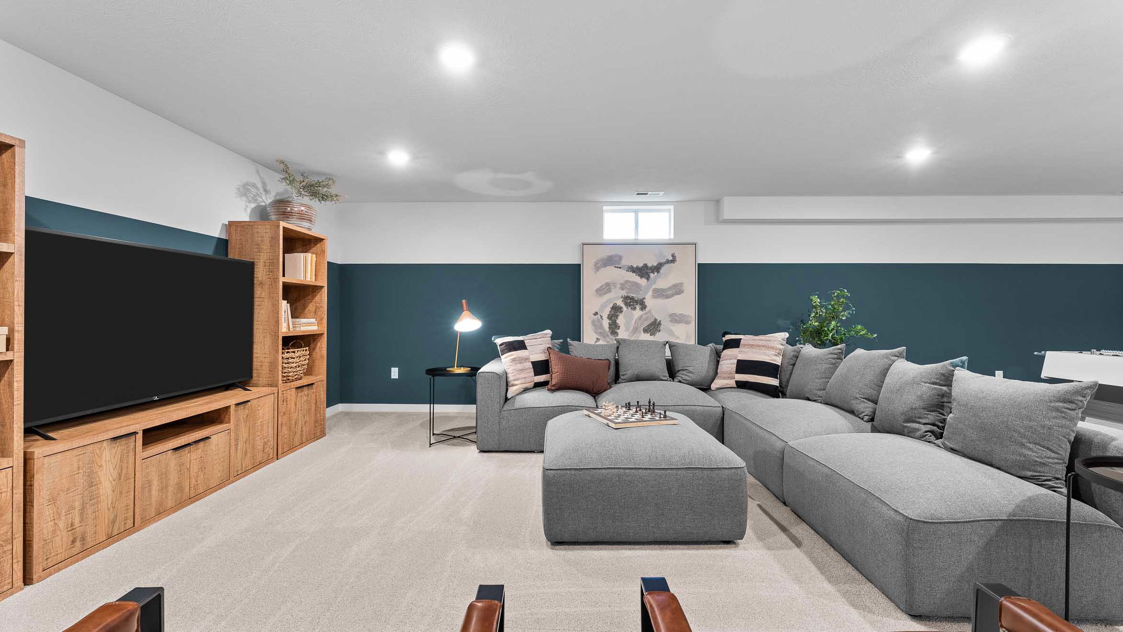 Modern living room with a large gray sectional sofa, wooden entertainment center, and decorative accents, such as a chessboard and framed art, set against a dark teal and white wall.