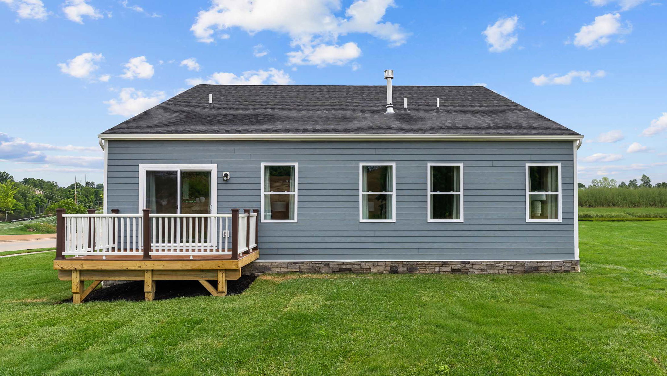 A modern, single-story blue house with a small wooden deck and a lush green lawn under a clear blue sky.