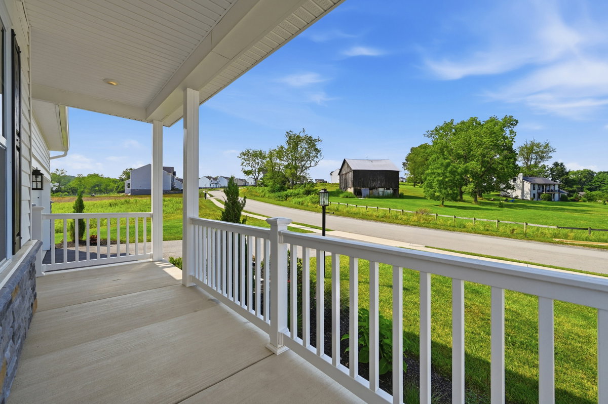 A view from a porch showing a grassy landscape with a road and distant barns under a clear blue sky.