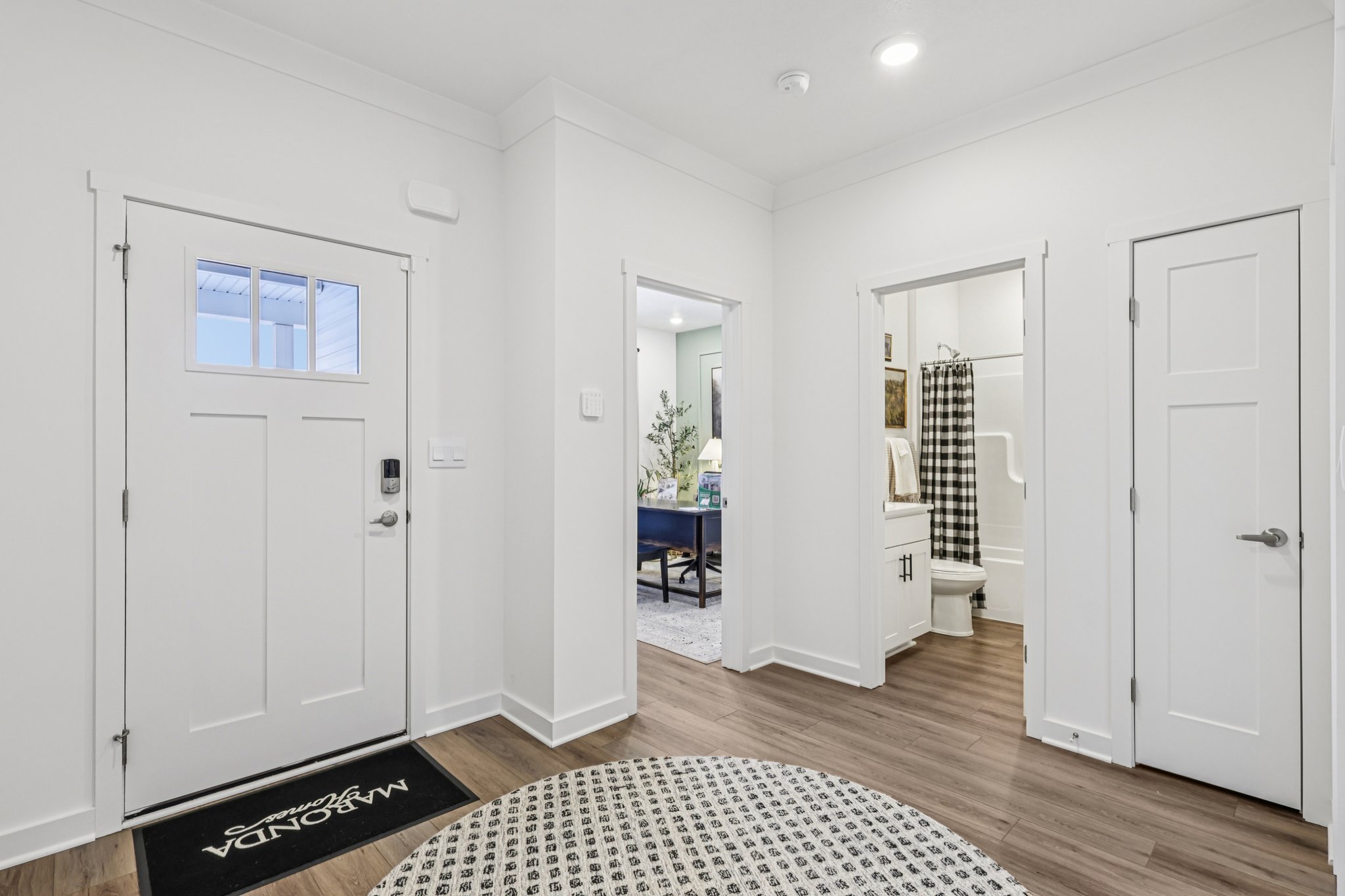 Modern entryway with a round patterned rug, open doors leading to a bathroom and a living area, and bright white walls.