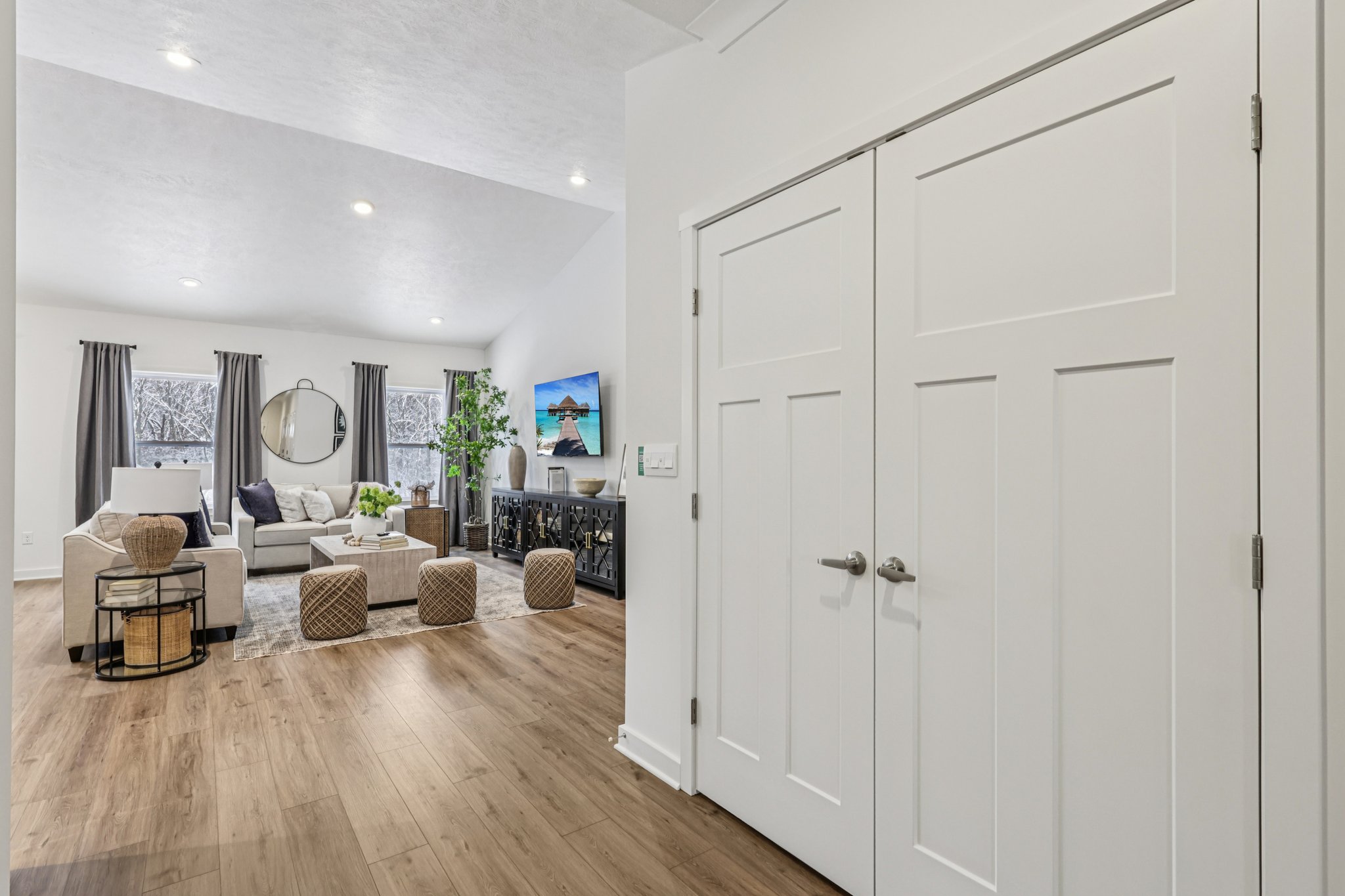 Entryway leading to a modern living room with neutral-colored furniture, a round mirror, and a large wall-mounted television.
