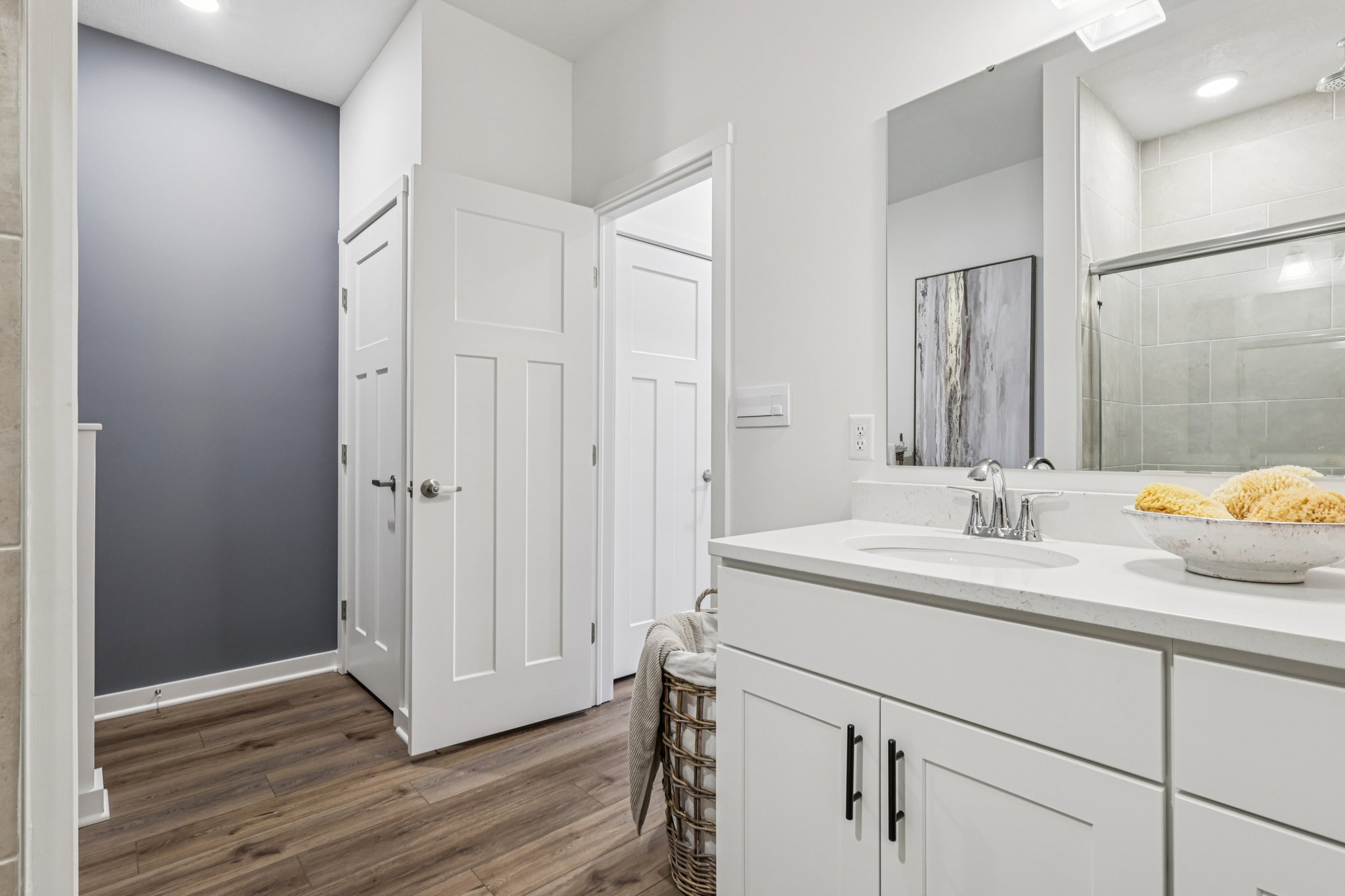 A modern bathroom featuring white cabinetry, a sink, and a shower, with a hallway and two doors visible in the background.