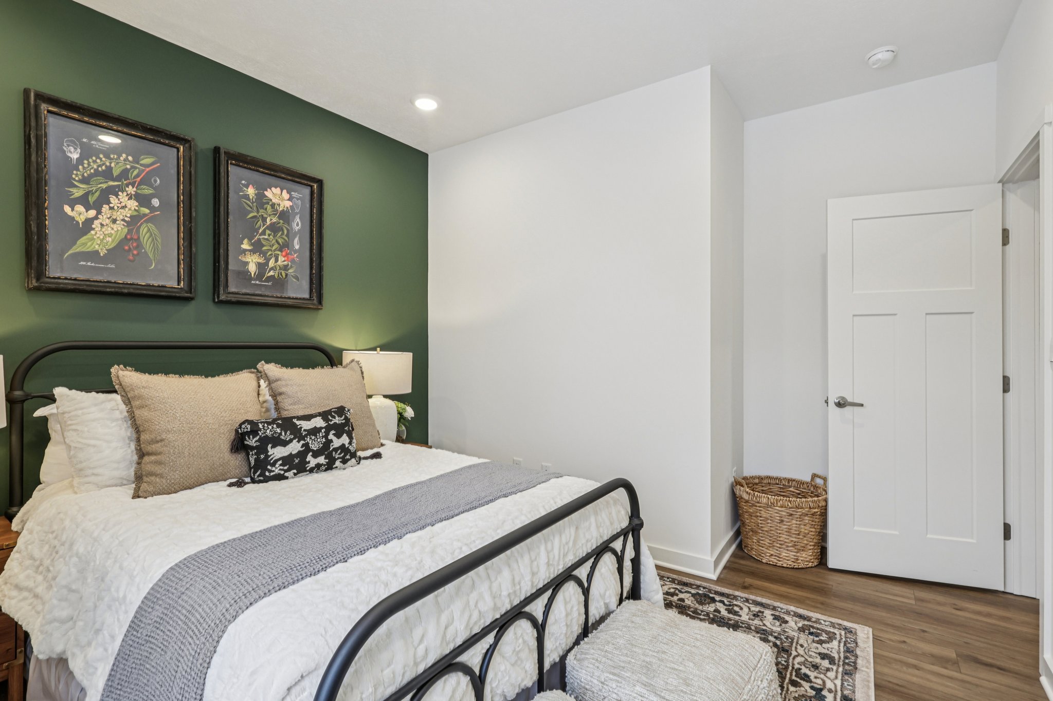 Cozy bedroom featuring a black metal bed, decorative pillows, green accent wall, and two framed botanical prints.