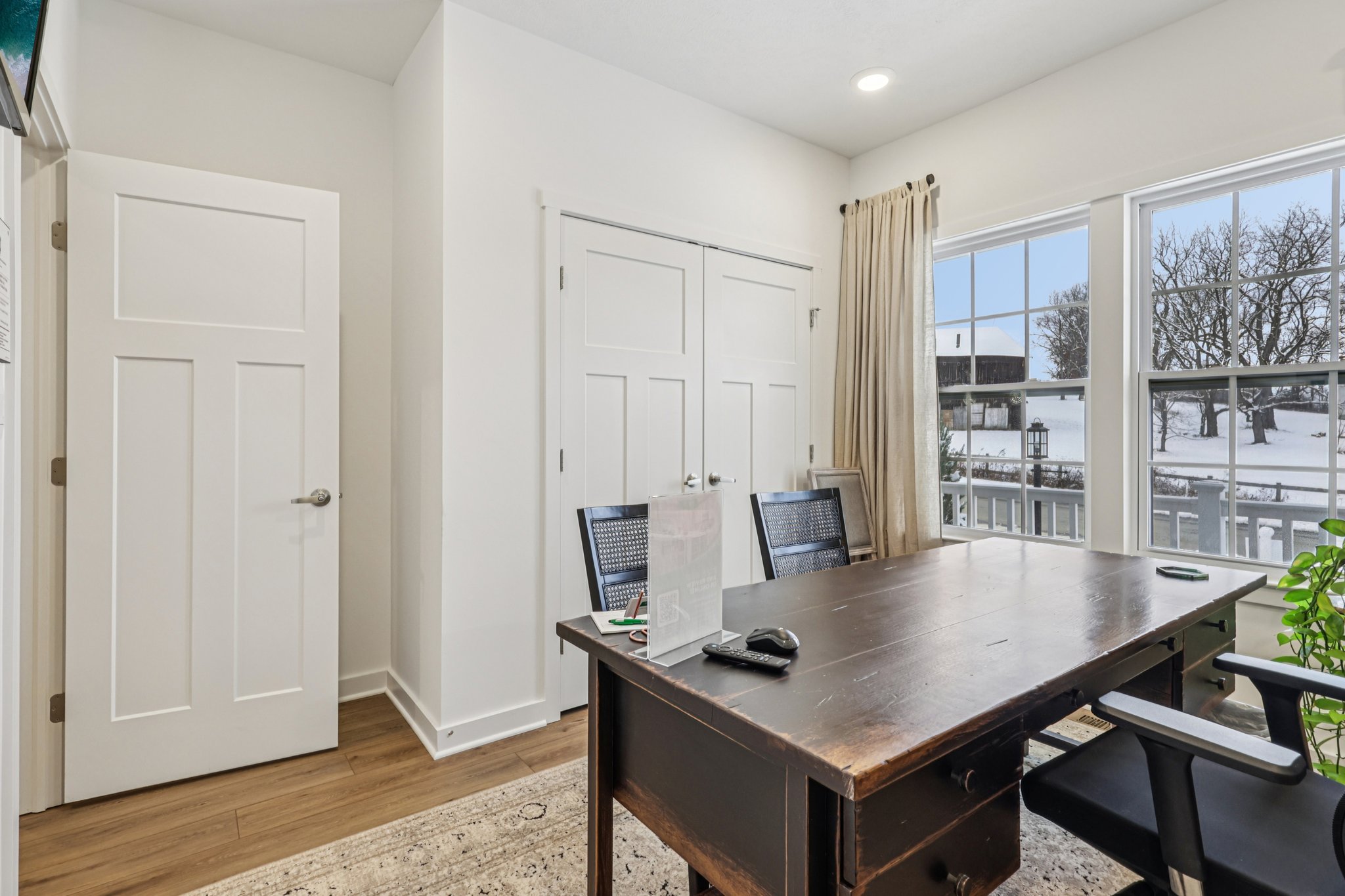 A home office with a wooden desk, two chairs, and windows showing a snow-covered landscape outside.