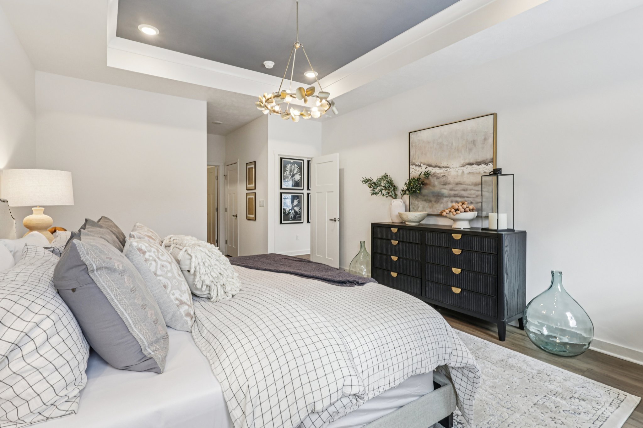 A stylish bedroom featuring a bed with gray and white bedding, a black dresser, decorative wall art, and a large vase.
