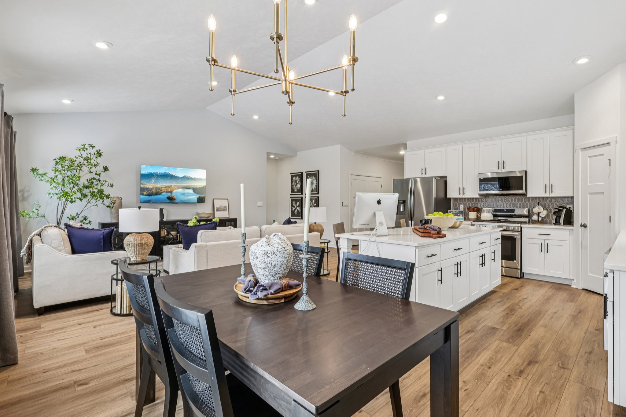 Modern kitchen and dining area with a wooden table, black chairs, and a bright living space featuring a television and greenery.