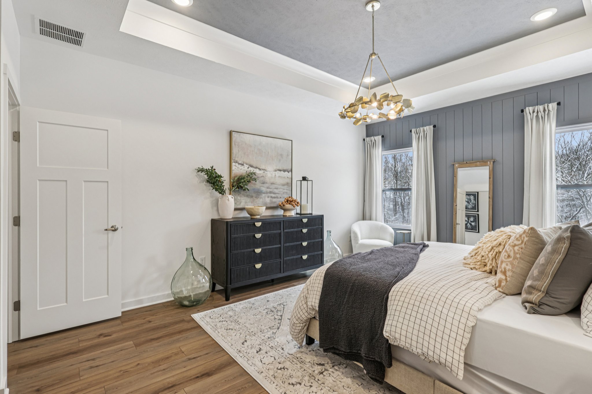 A stylish bedroom featuring a gray accent wall, a large bed with textured blankets, a modern dresser, and decorative elements like a painting and plants.