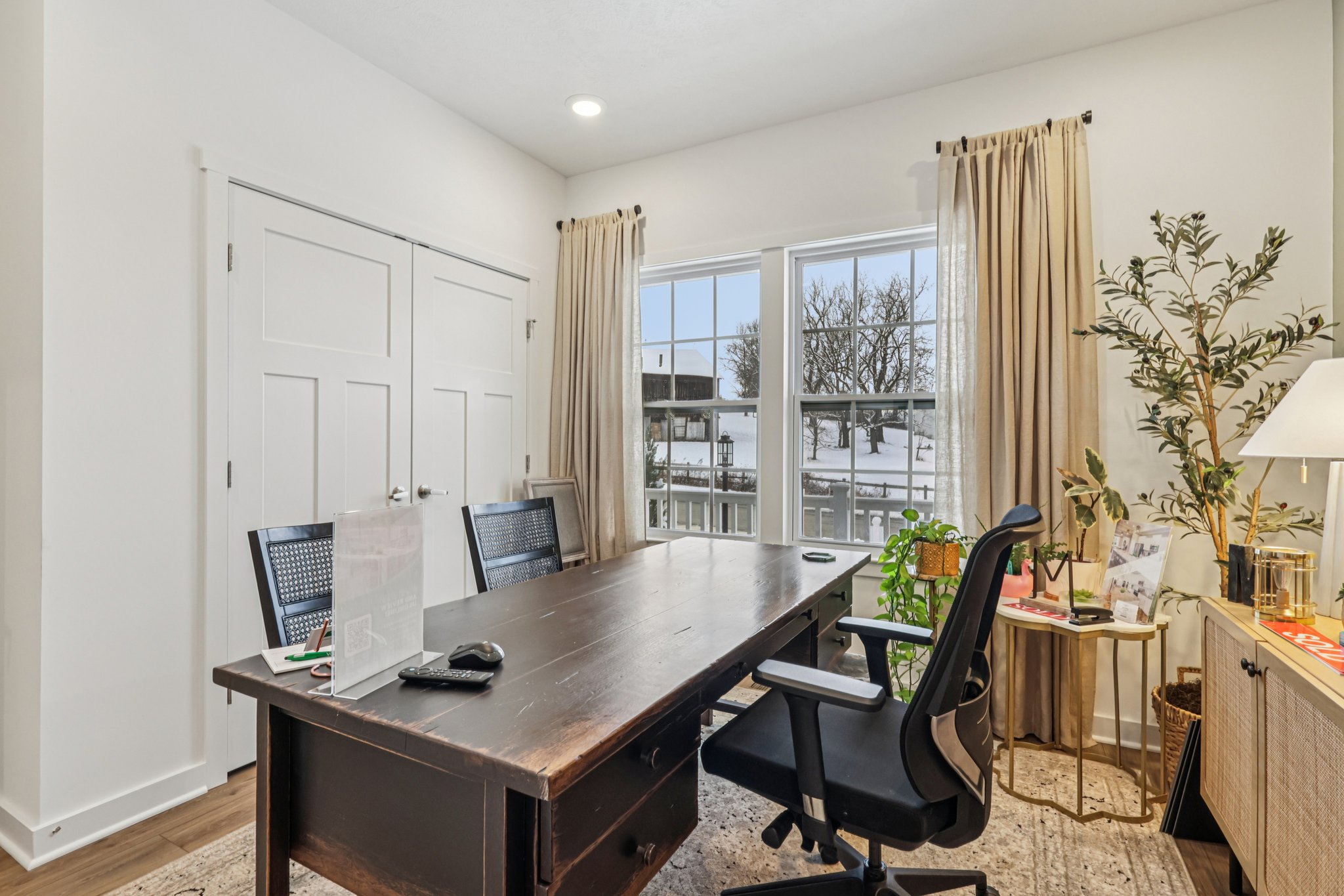 A modern home office featuring a large wooden desk, black chairs, and natural light streaming through window curtains.