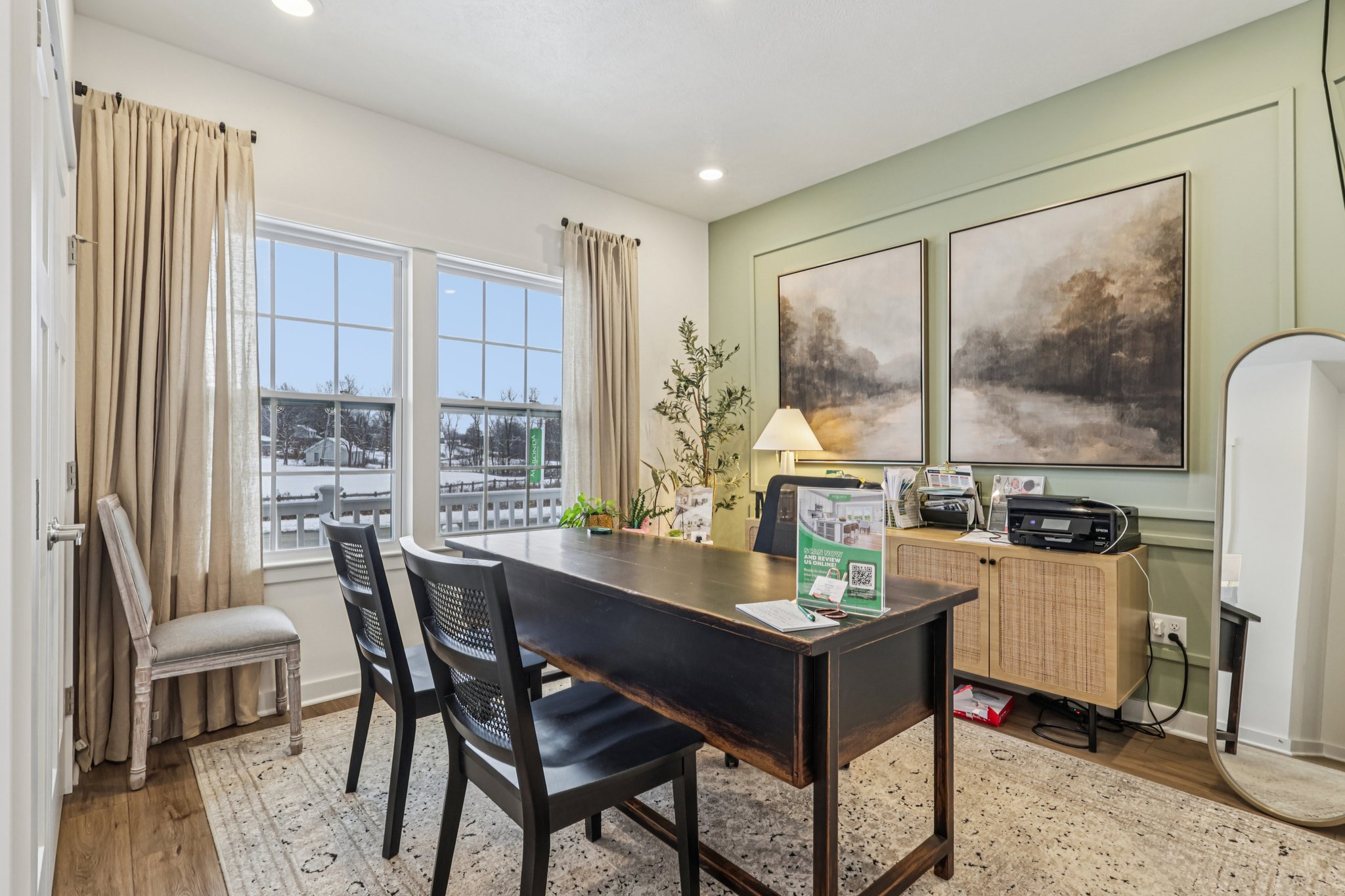 A cozy home office features a dark wooden desk, two black chairs, large windows with beige curtains, and decorative artwork on the walls.