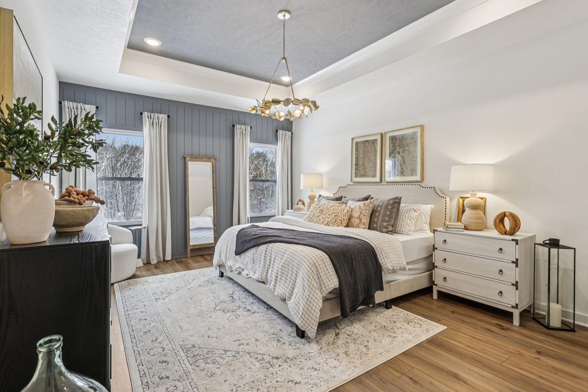 A modern bedroom featuring a bed with decorative pillows, a pair of bedside tables with lamps, a large mirror, and a cozy chair by the window.