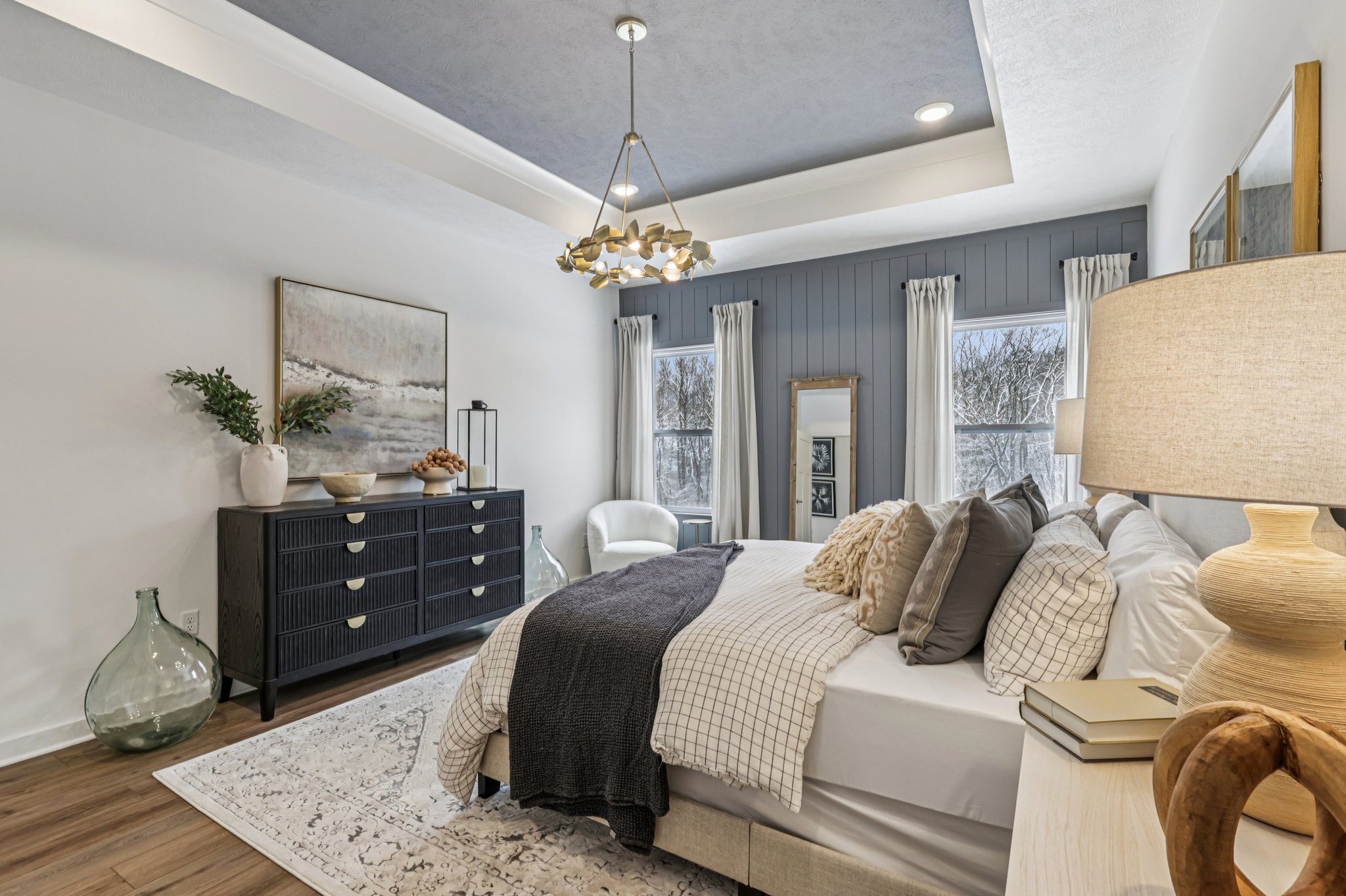 A modern bedroom featuring a bed with decorative pillows, a stylish nightstand, a large mirror, and a beige rug, illuminated by a chandelier.