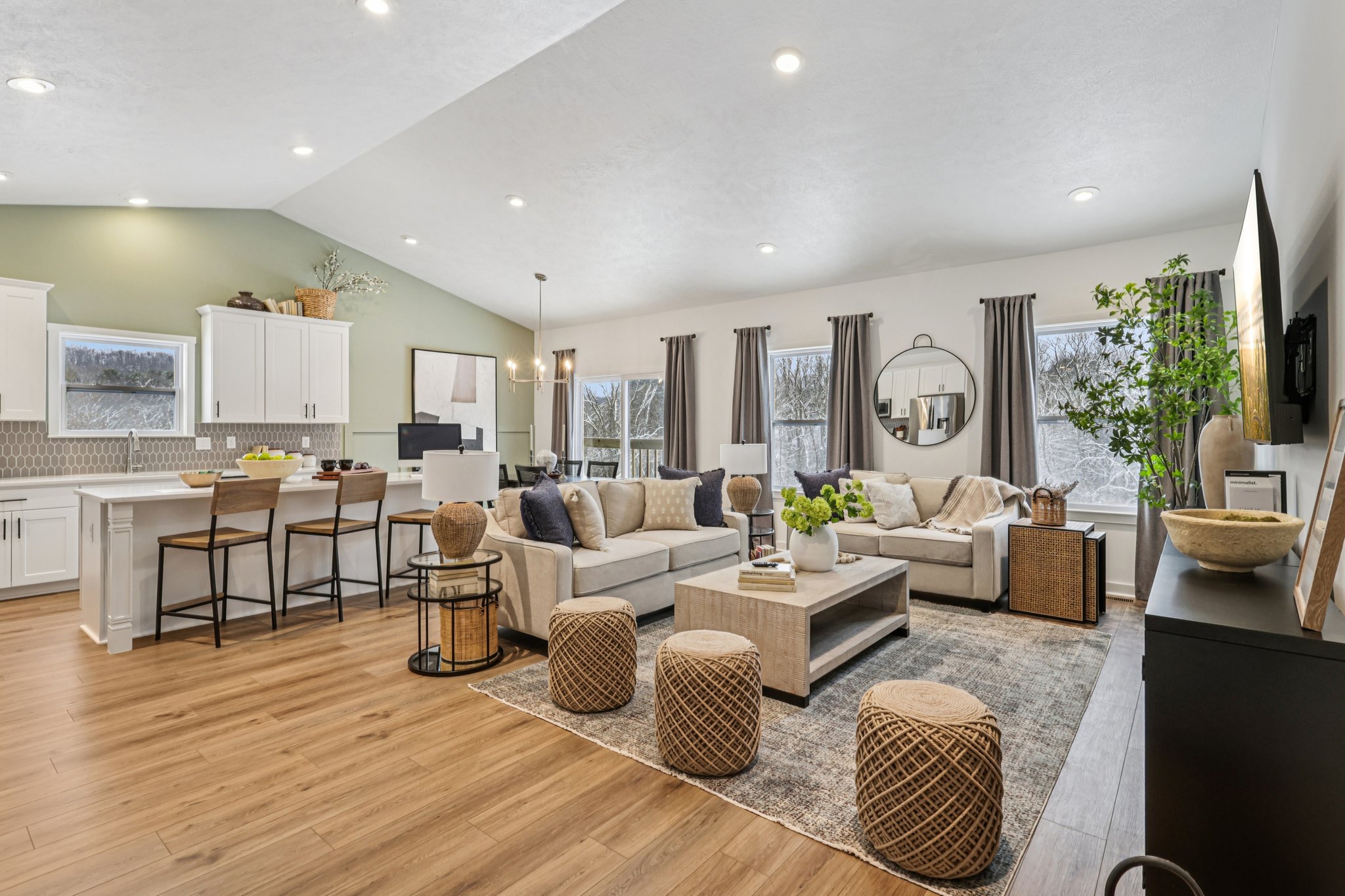 A modern living room featuring a light-colored sofa, wooden coffee table, and large windows, with a kitchen area visible in the background.
