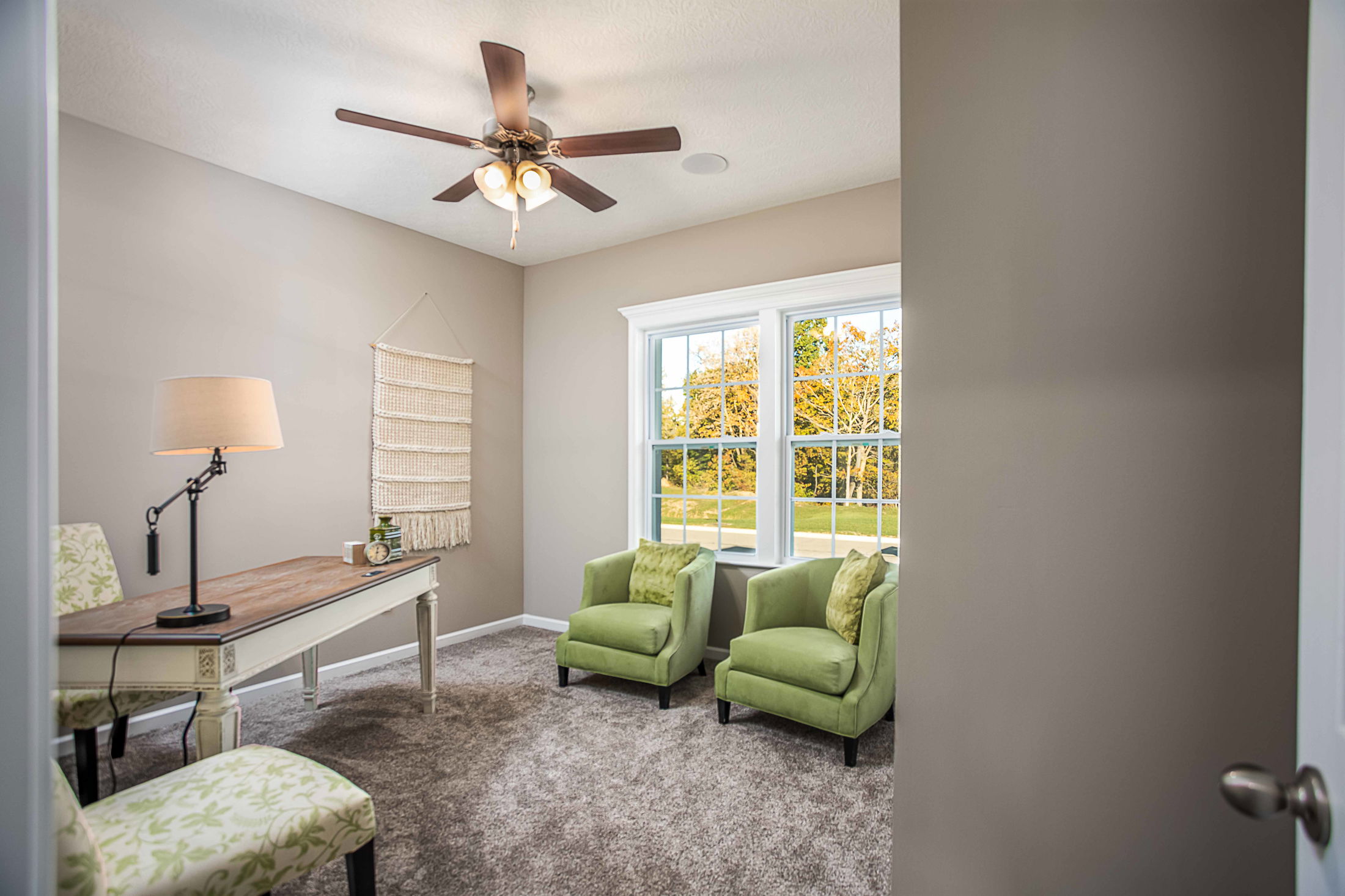 Cozy home office interior with green armchairs, a wooden desk, and a ceiling fan.