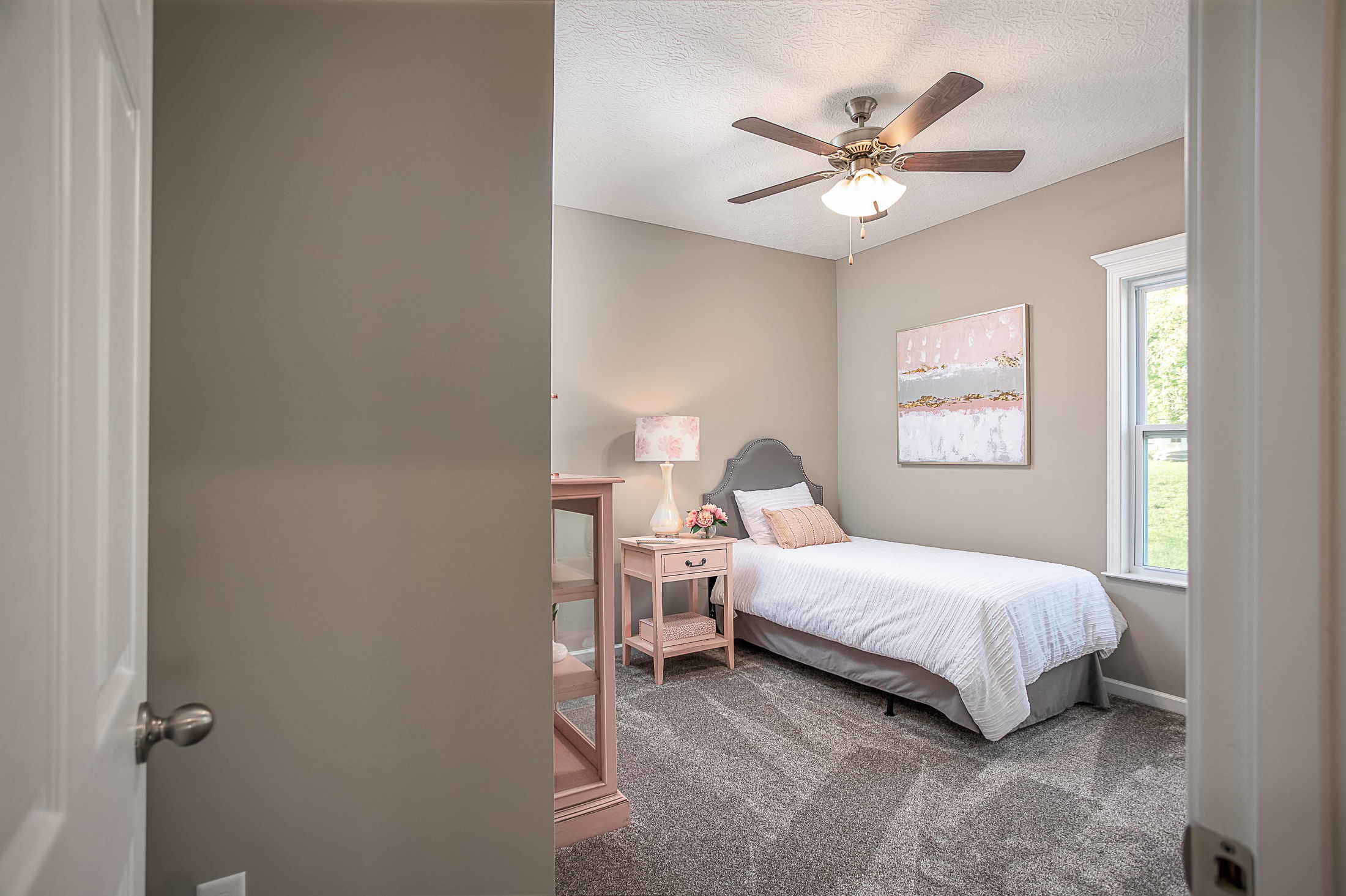 Cozy bedroom with pastel decor featuring a twin bed, nightstand, wall art, and ceiling fan, viewed through an open door.