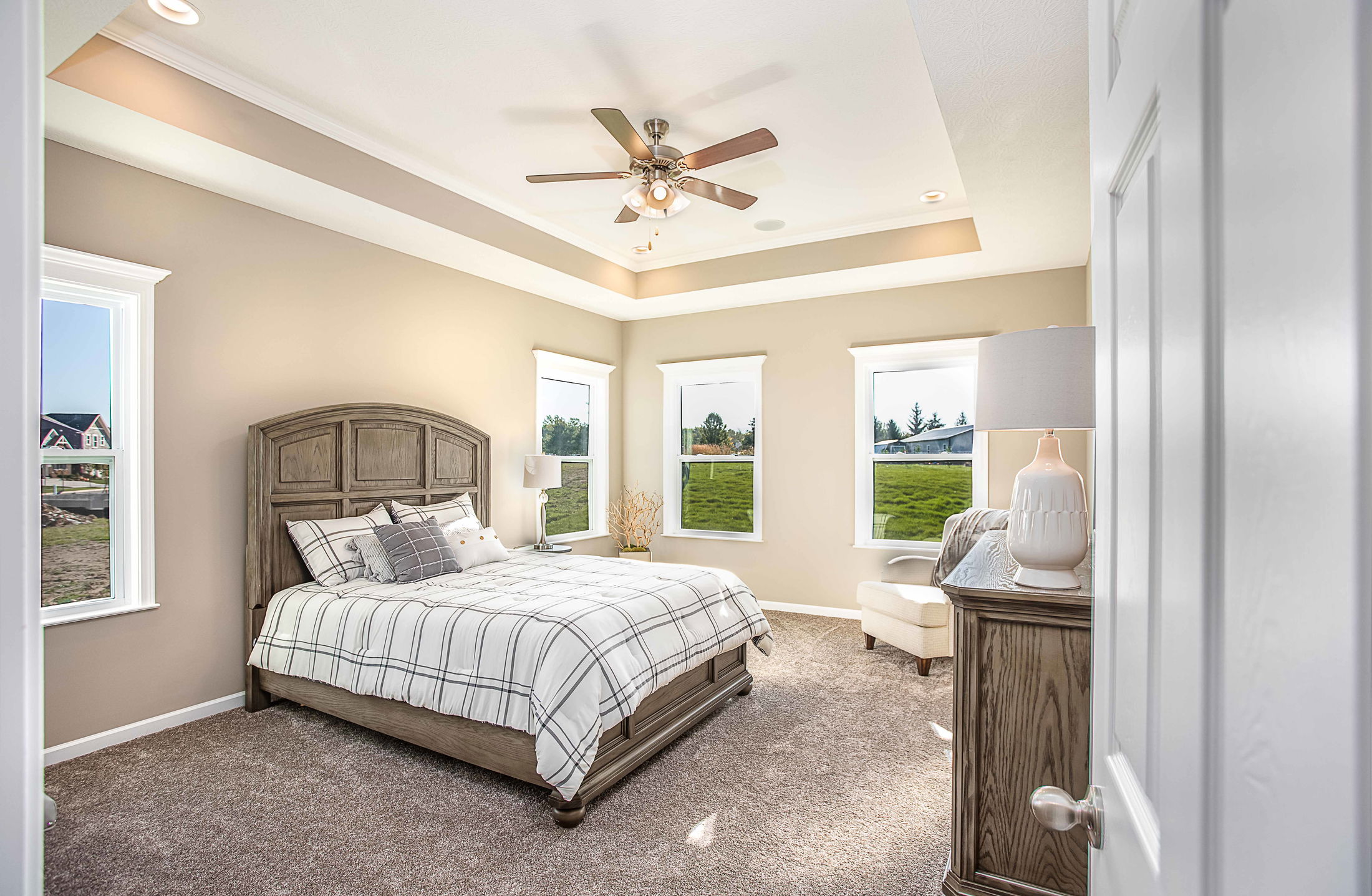 Elegant, sunlit bedroom featuring a wooden bed with plaid bedding, a ceiling fan, carpeted floor, and modern decor.