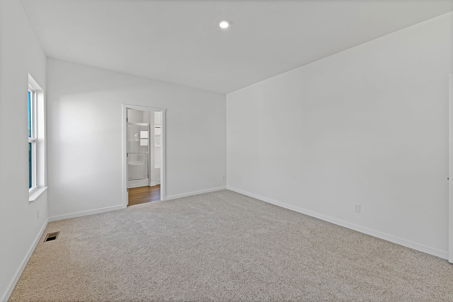 A bright, empty room with white walls, beige carpet, and a doorway leading to a bathroom with a glass shower.