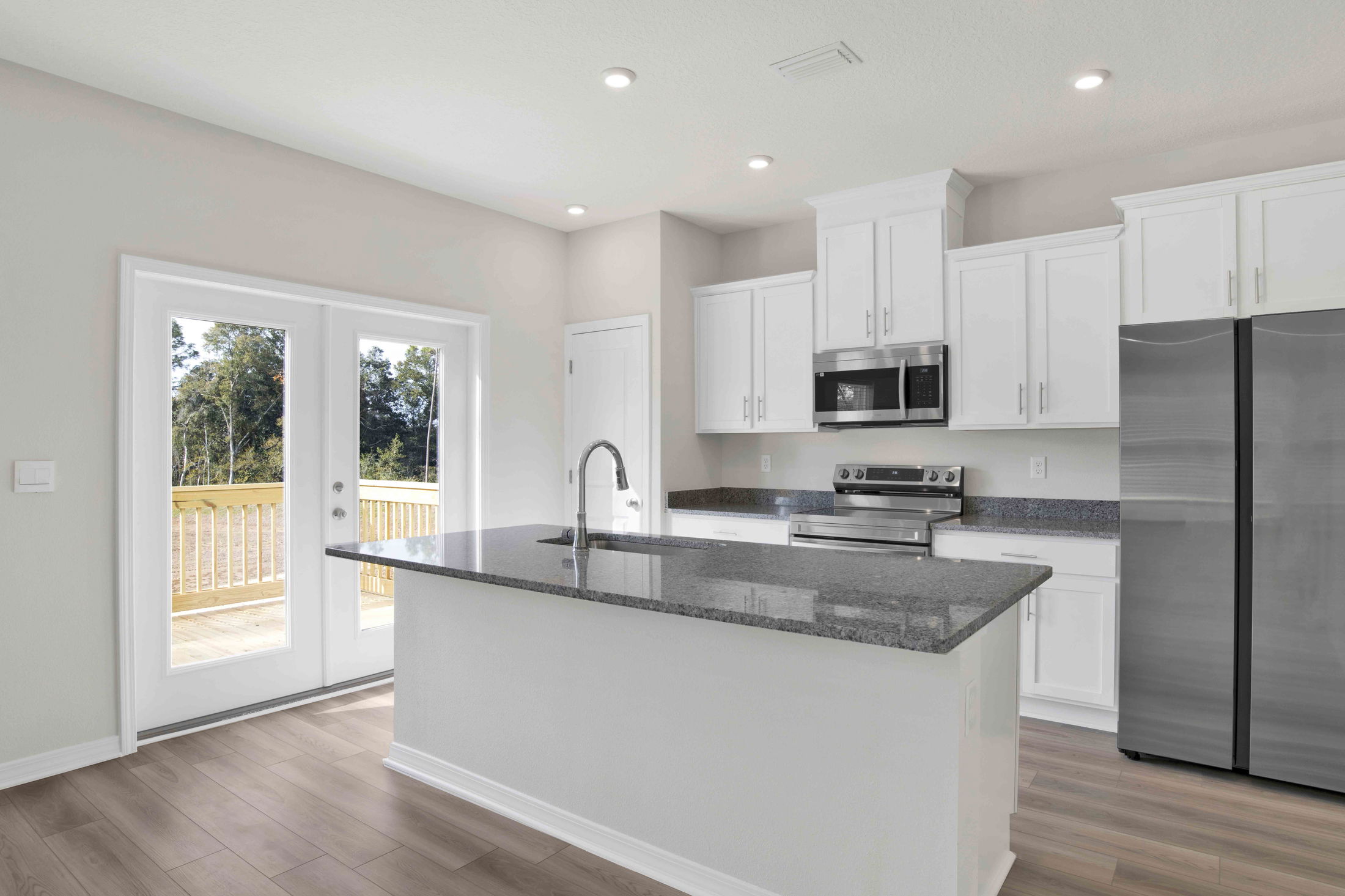 Modern kitchen with white cabinets, granite countertops, stainless steel appliances, and a center island, overlooking a wooden deck through French doors.
