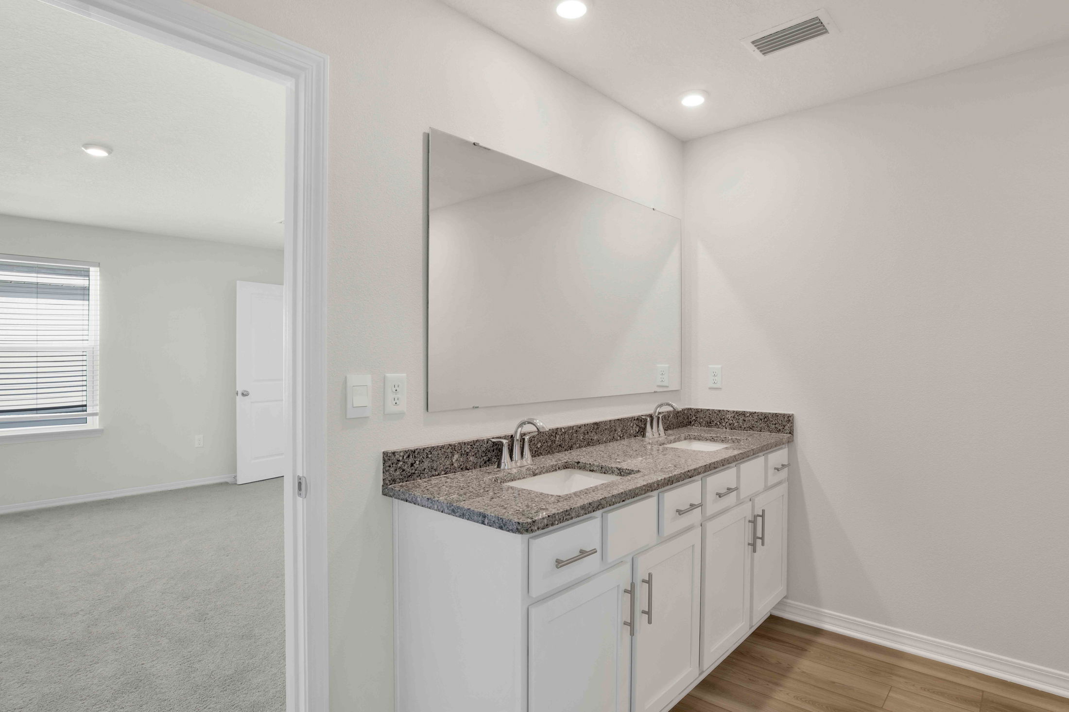 Modern bathroom with a double sink vanity and granite countertop next to a bright carpeted bedroom.