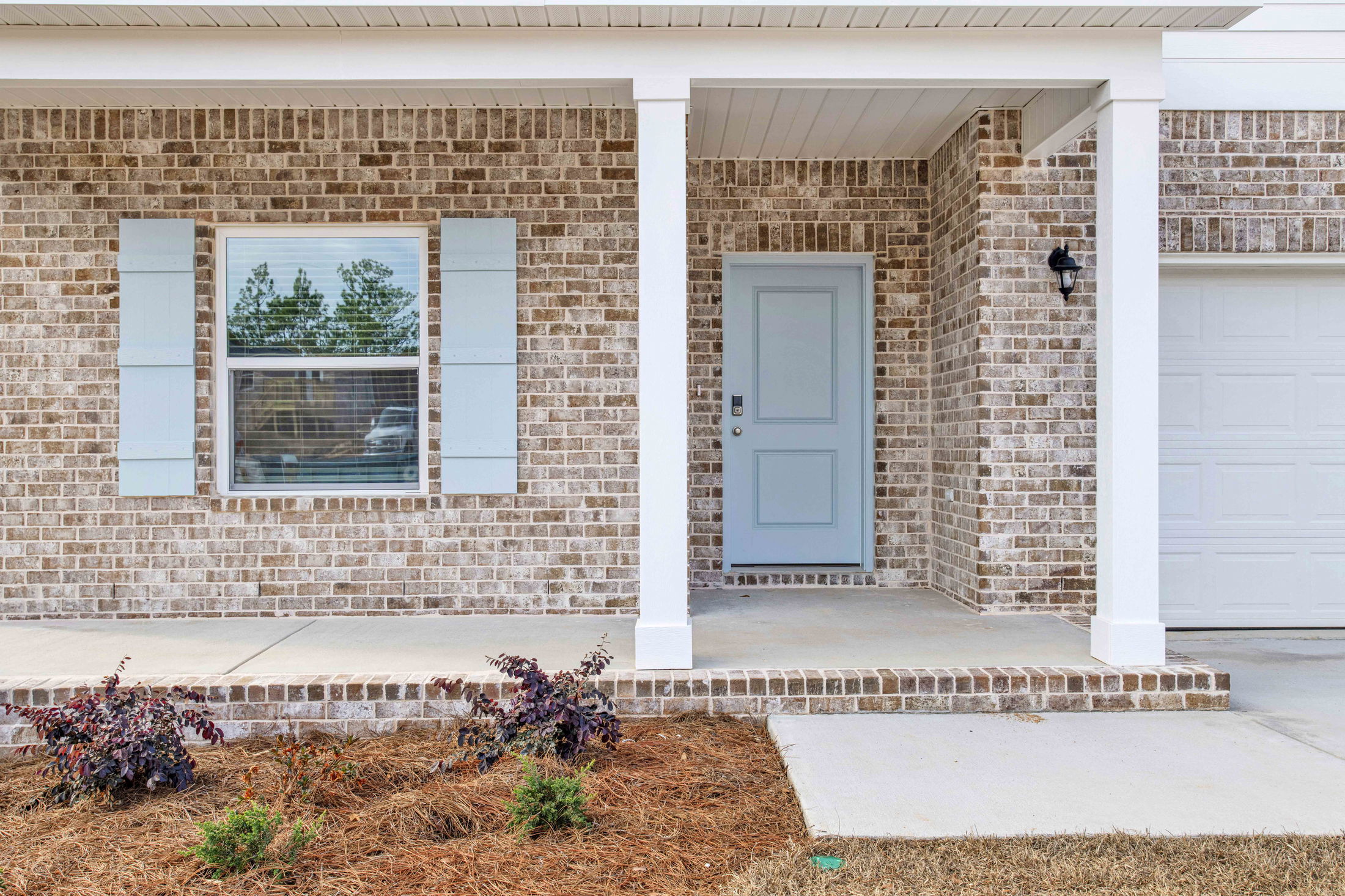 Front entrance of a modern brick home with a light blue door, white shutters, and manicured landscaping.