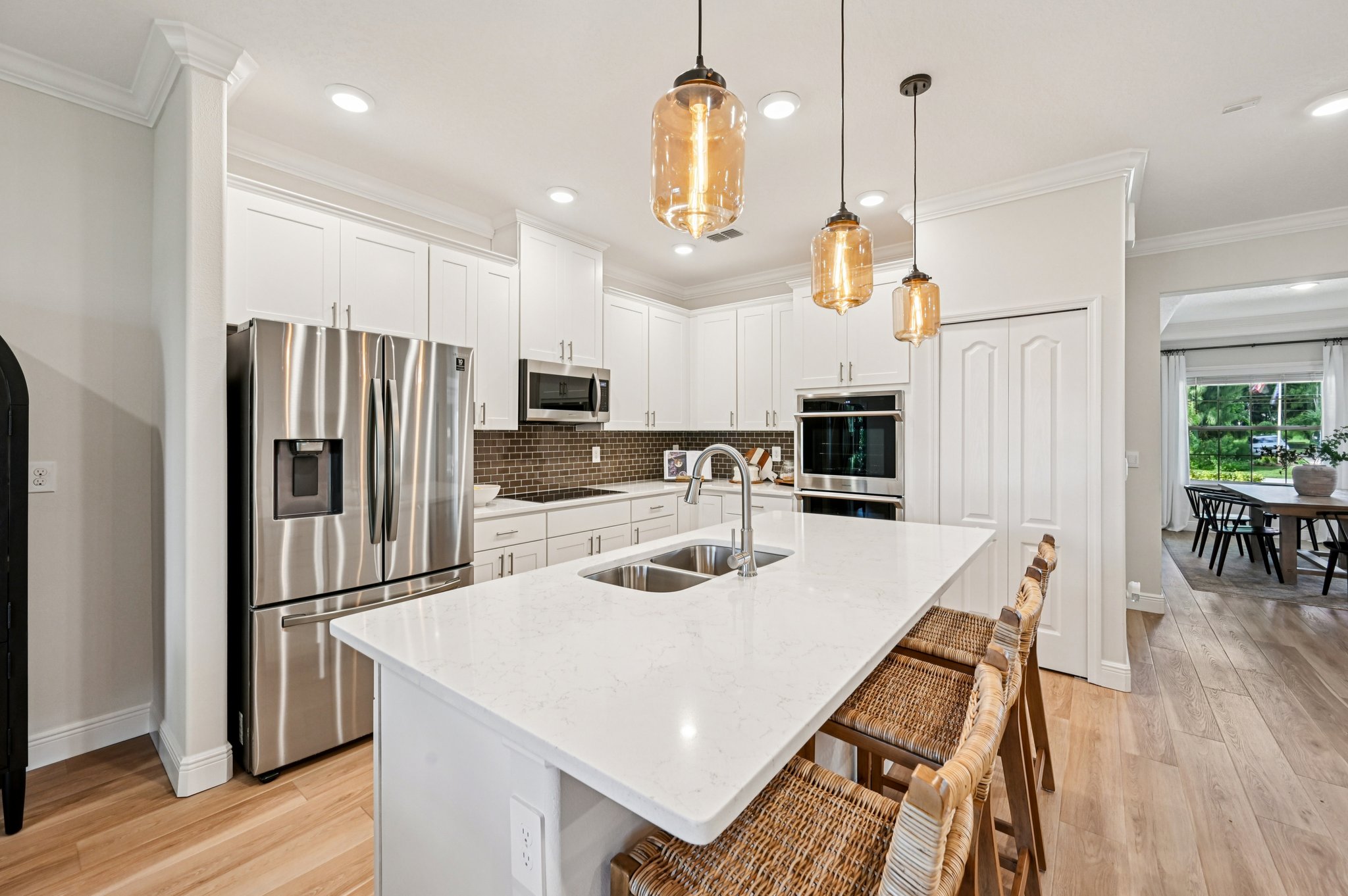 Modern kitchen with a sleek white island countertop, stainless steel appliances, wicker bar stools, and pendant lighting.