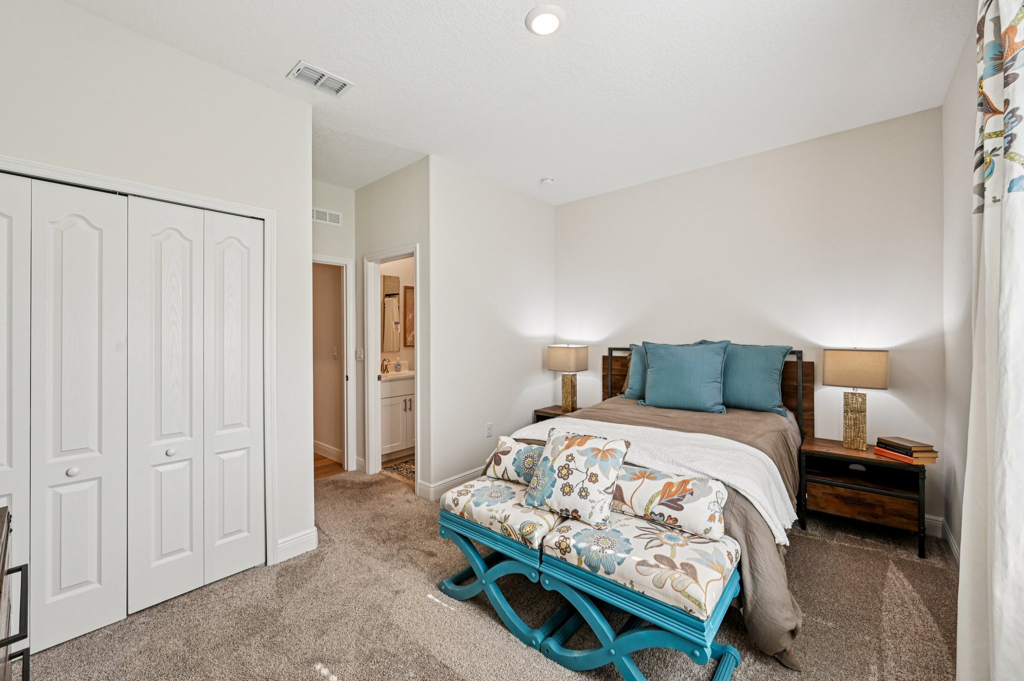 Elegant bedroom with a cozy bed, blue and floral decor, carpeted floor, and a glimpse into an adjacent bathroom.