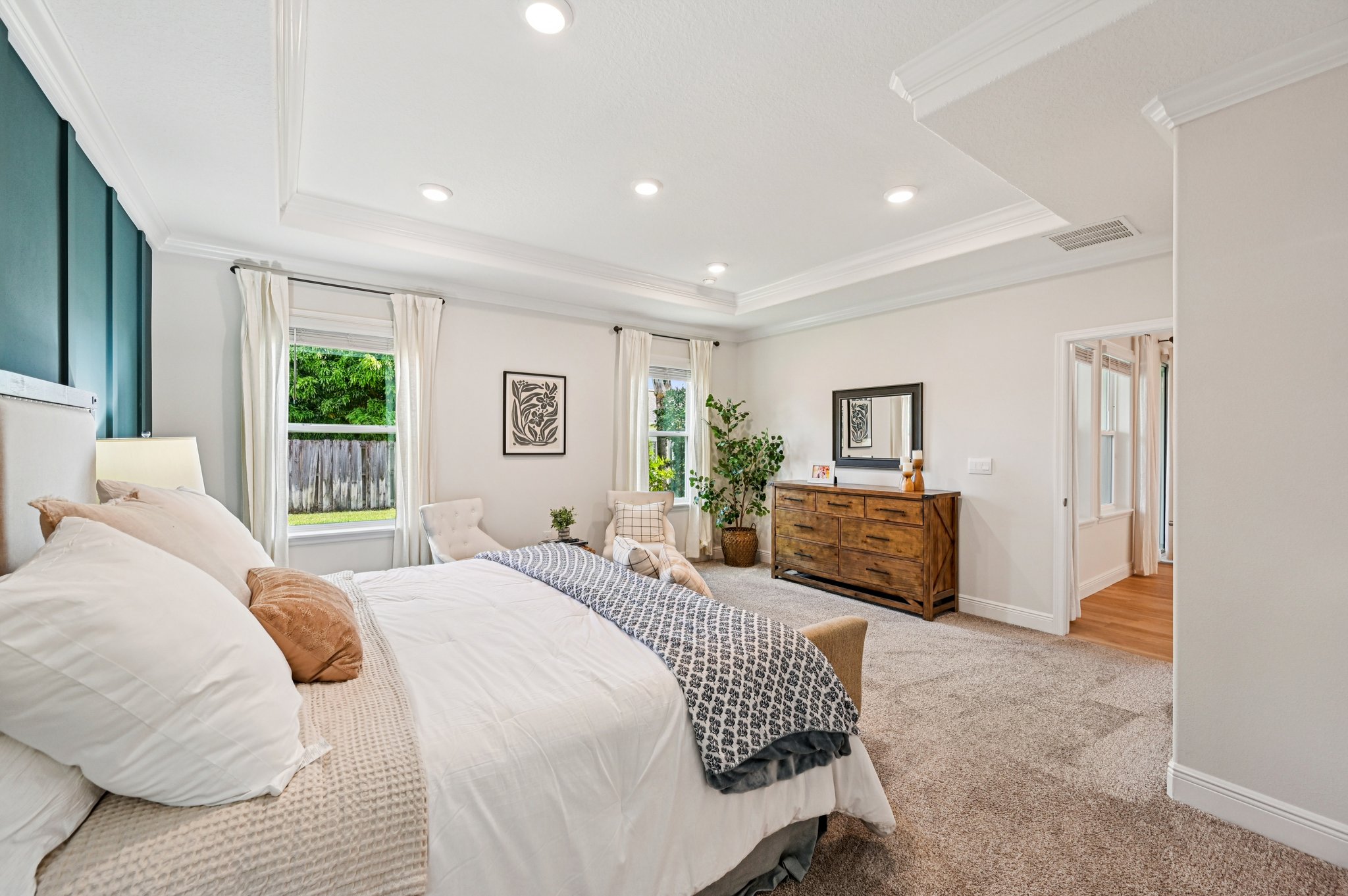 Modern master bedroom with plush carpet, white bedding, wooden dresser, and large windows offering natural light.