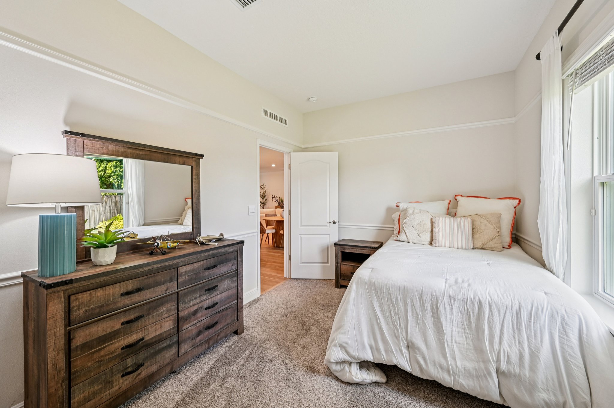 Cozy bedroom with white bedding, wooden dresser, and large window letting in natural light.