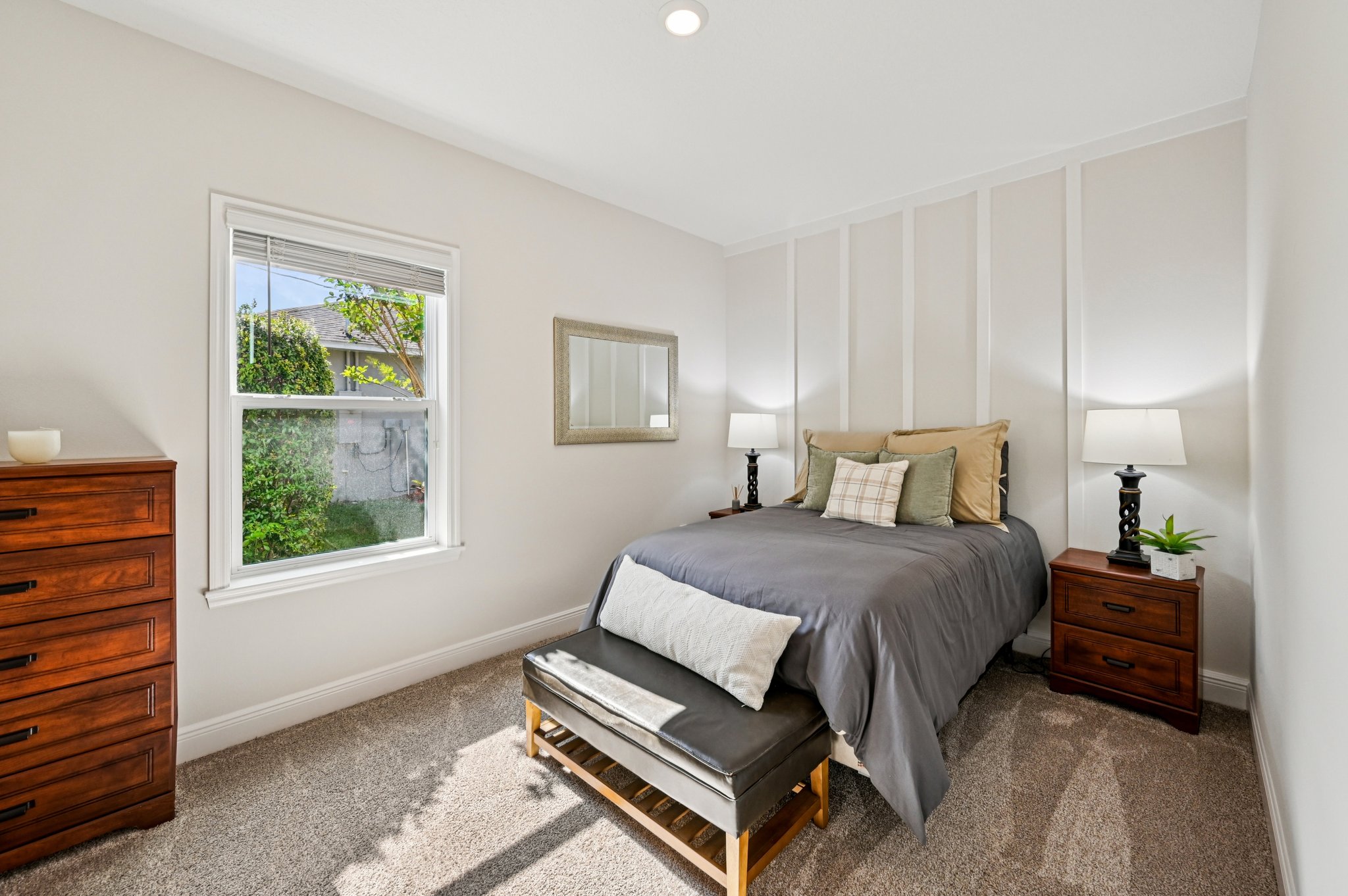 Bright and cozy bedroom with a neatly made bed, accent pillows, wooden furniture, and a window overlooking greenery.