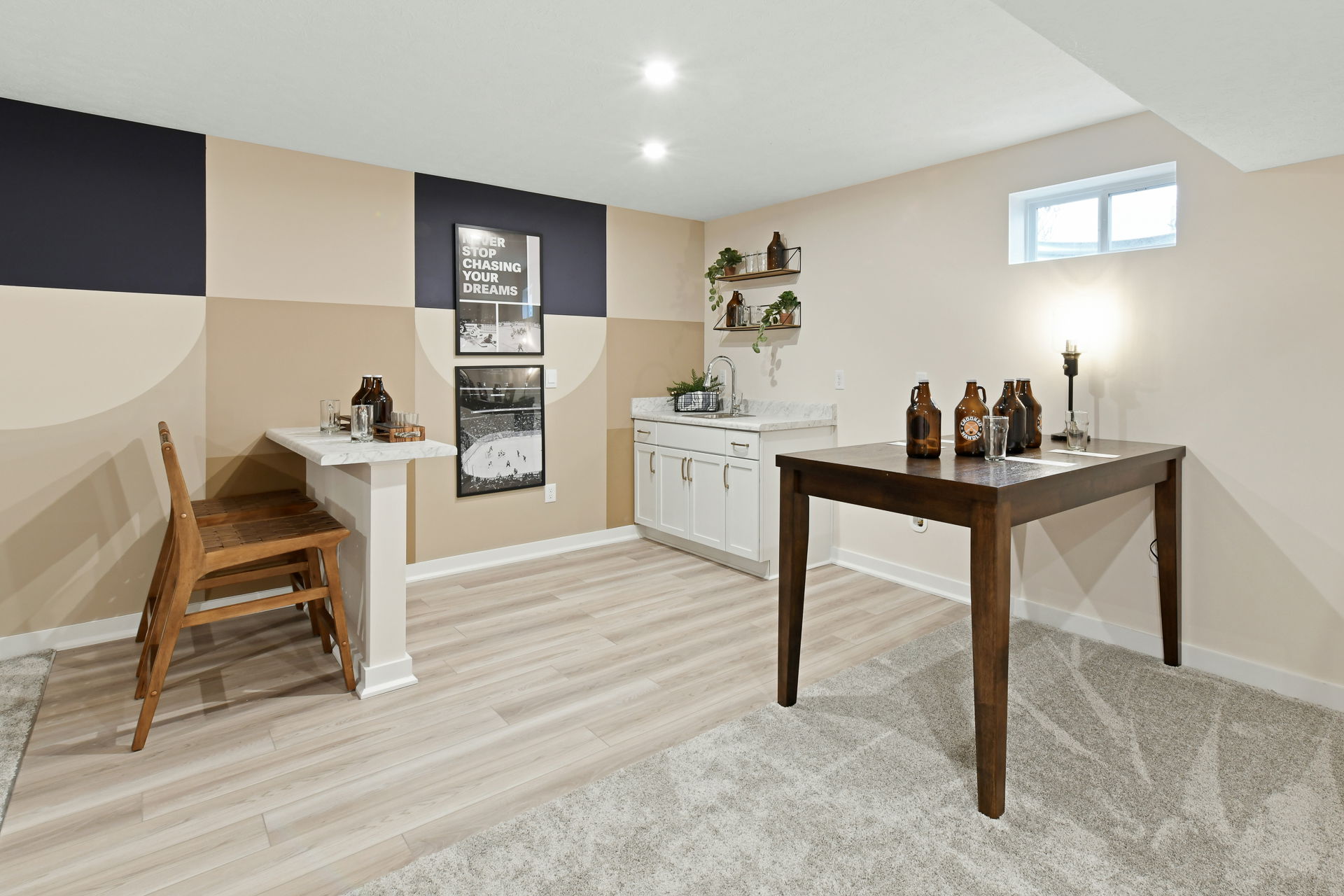 Modern basement bar area with a beige and navy accent wall, featuring a small wooden table, bar stools, and stylish shelving.