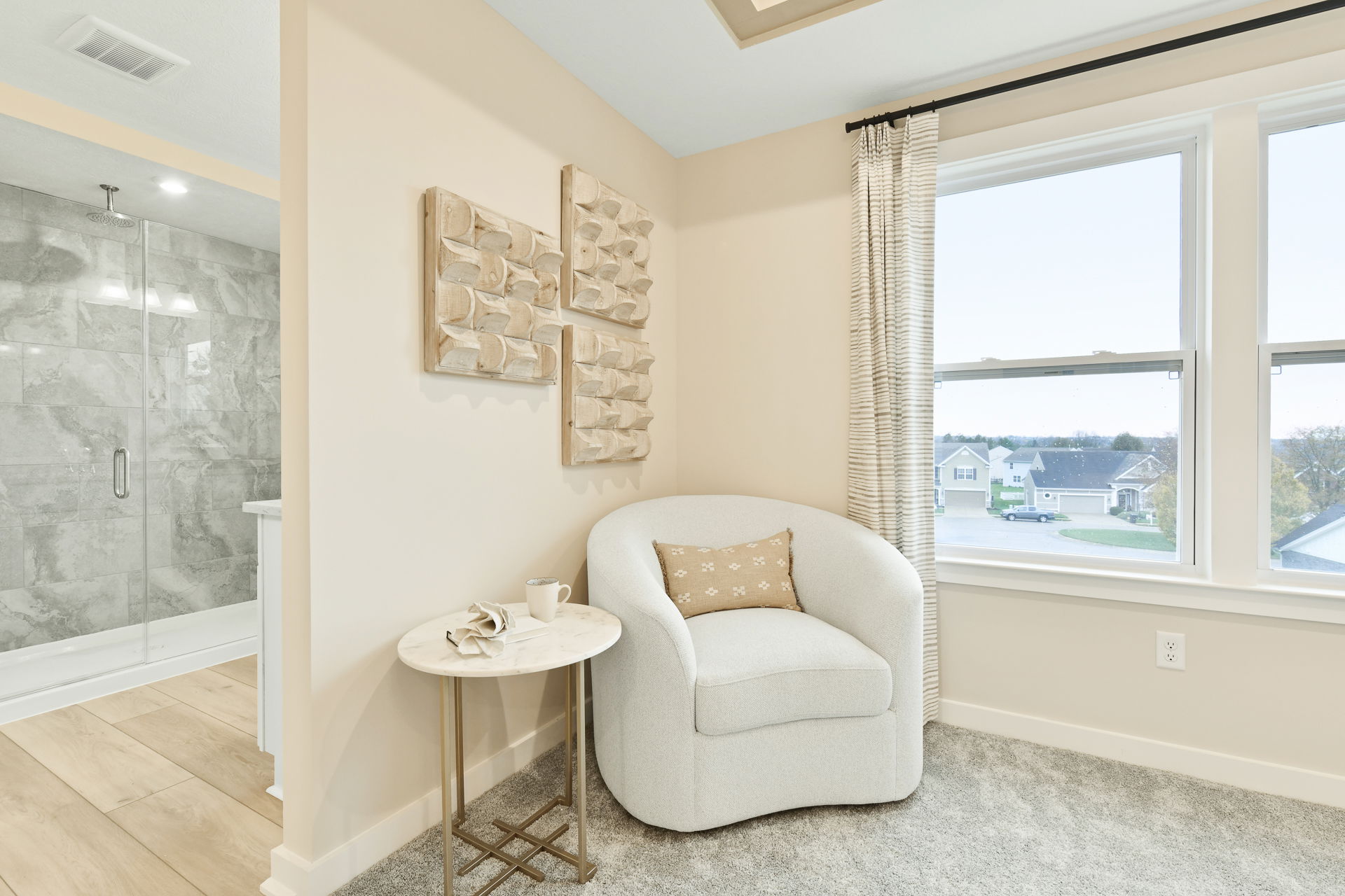 Cozy modern living room corner with a white armchair, decorative pillow, round marble side table, abstract wall art, and large windows overlooking a scenic neighborhood.