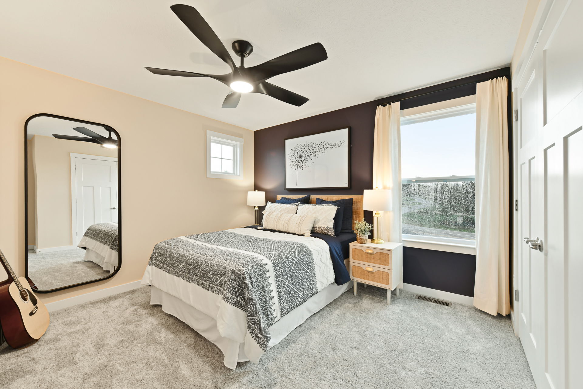 A modern bedroom featuring a cozy bed with a geometric patterned comforter, large black ceiling fan, full-length mirror, and a guitar on the carpeted floor.