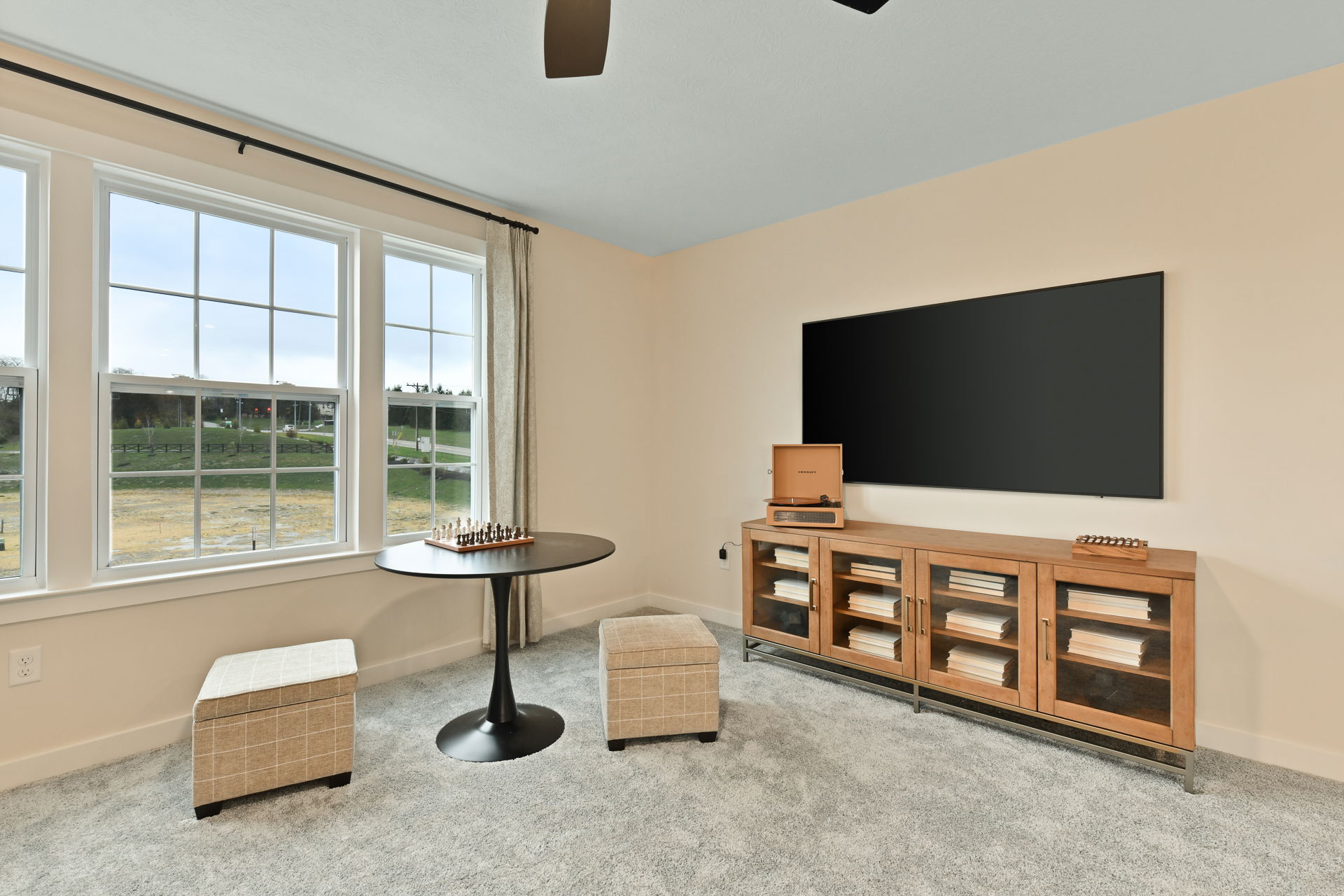 A cozy living room with a large window, round table with a chess set, cushioned stools, and a wooden cabinet beneath a wall-mounted TV.